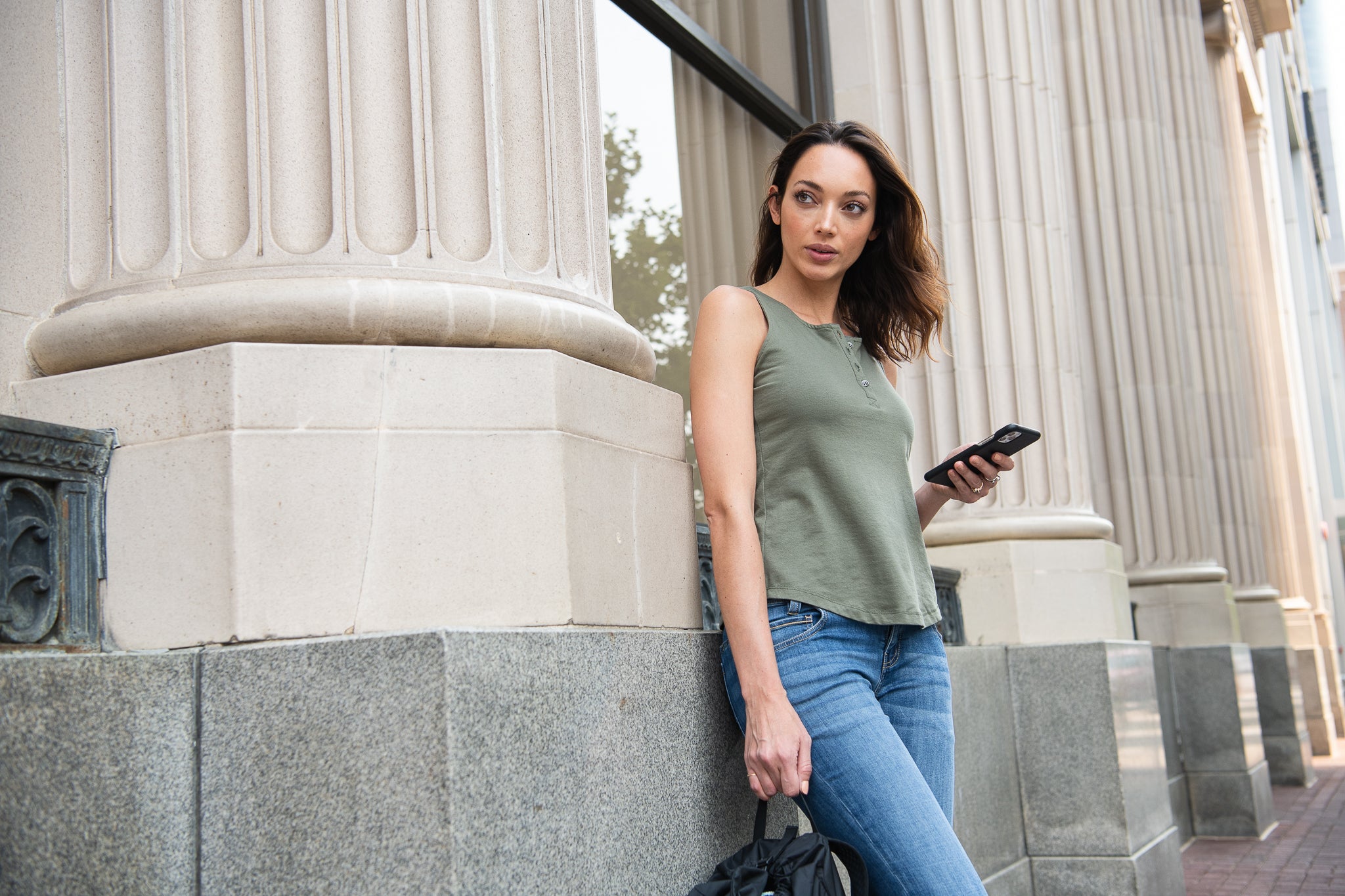 Photograph of a woman in a sage green tank top and blue jeans holding a smartphone against a building with classical columns, a granite base, and ornate detailing