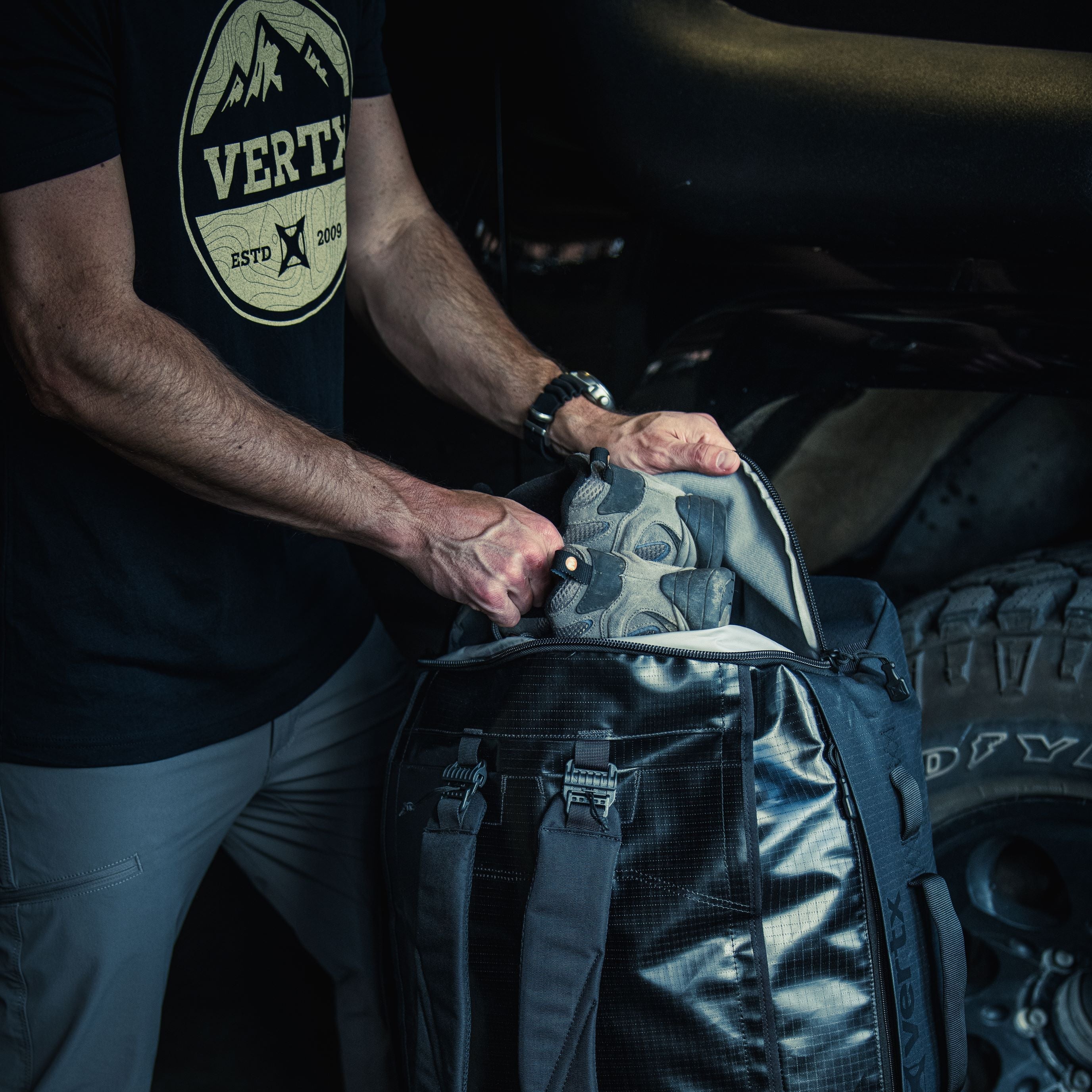 A photograph shows a person wearing a Vertyx t-shirt packing gray hiking boots into a black Vertyx backpack near a vehicle tire with a rugged tread