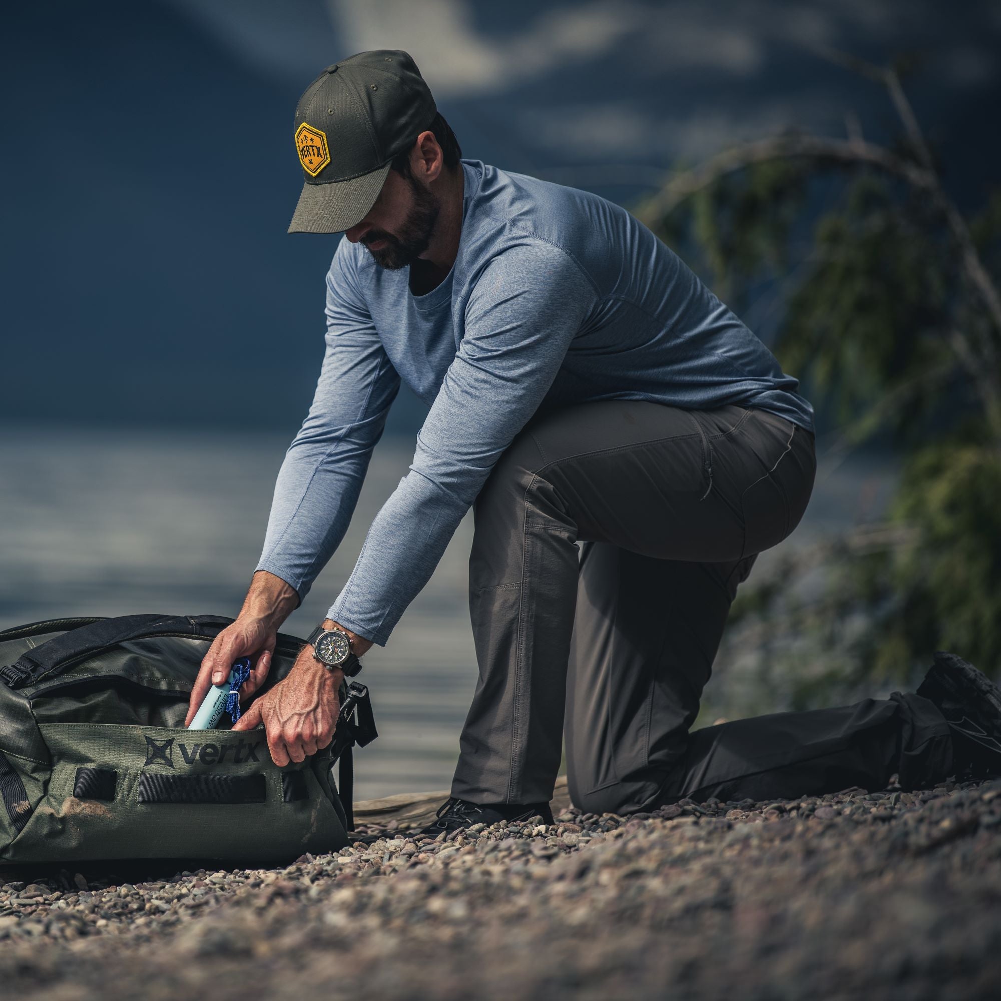 Photography, unknown artist: A man kneeling by a lake, packing a Vertx bag near rocky shoreline with mountains in the background, wearing a green cap with a logo, a blue shirt, and a detailed watch