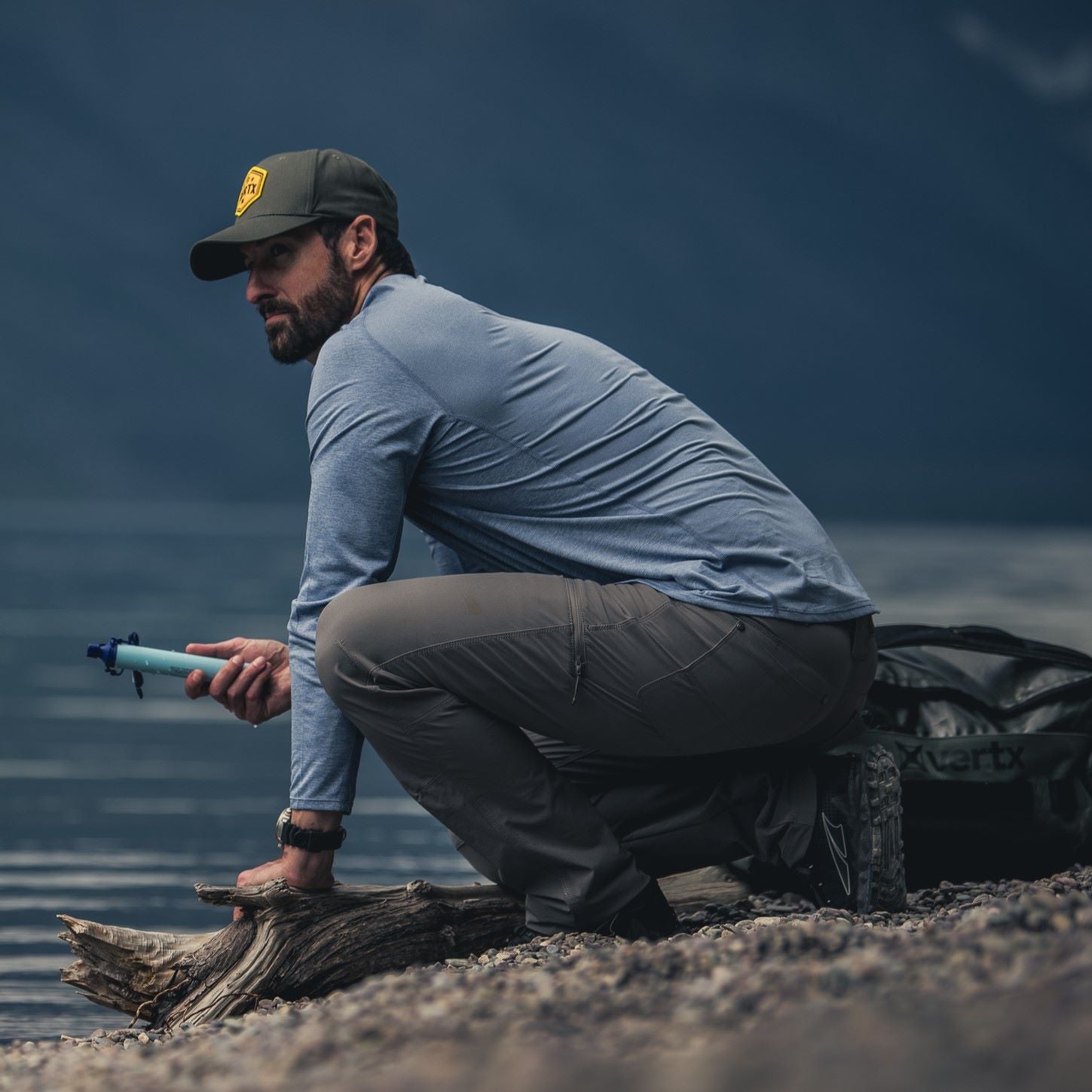 Photography Unknown Artist a man filters water near a lake using a blue filter with a weathered log and dark grey Vertix bag in the foreground against a dramatic mountain backdrop