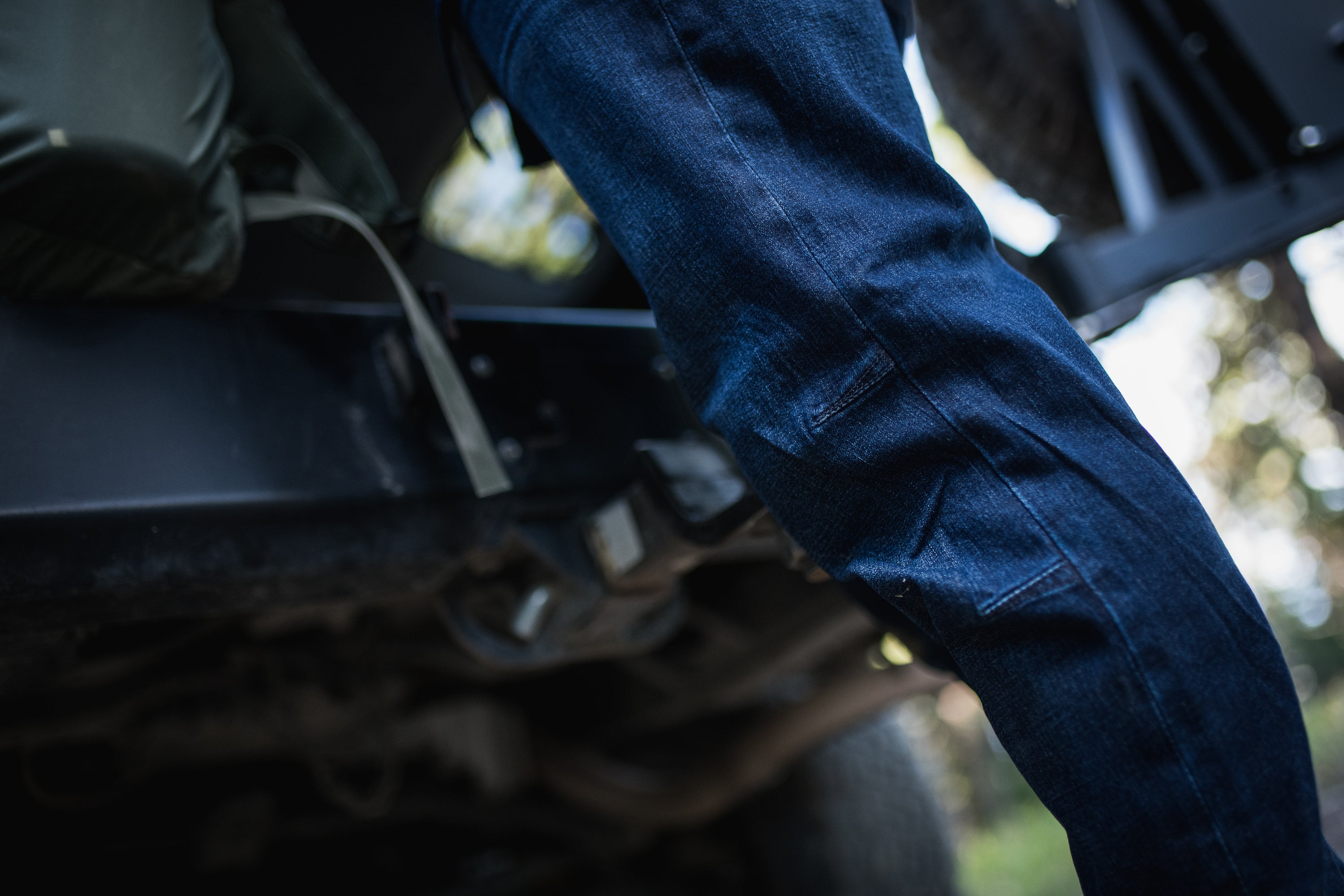 Photograph of dark wash denim jeans legs near a dark-colored vehicle in a forest showcasing the fabric texture, reinforced knee patches and subtle wear details