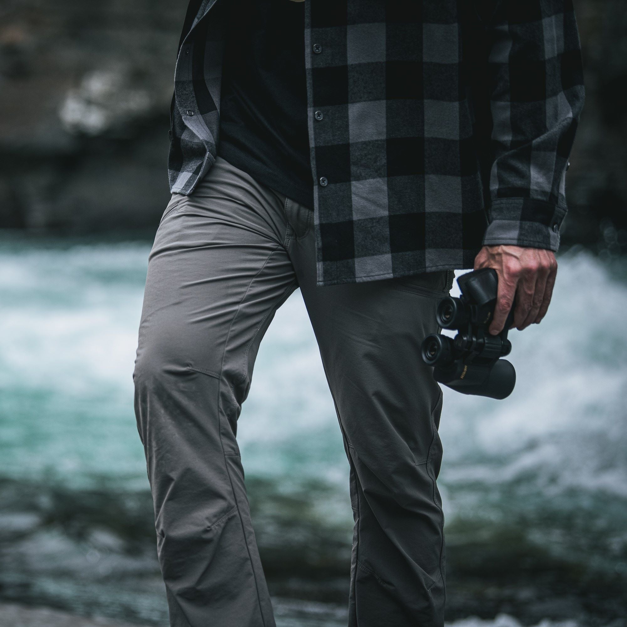 Photograph of a person wearing grey pants and a grey and black plaid shirt holding binoculars by a rushing river with dark rocks and blurred teal water.