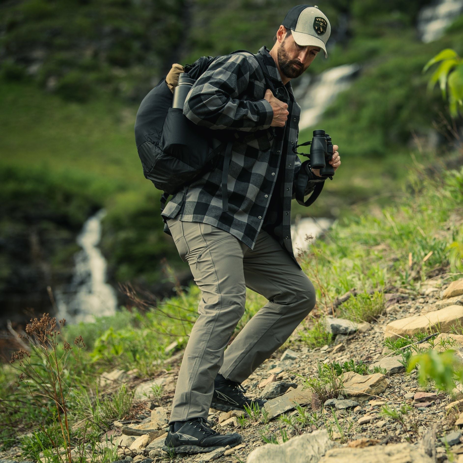 A photograph depicting a man hiking a rocky mountain trail near a waterfall, he is wearing a grey and black plaid shirt, olive green pants, and a baseball cap, carrying binoculars and a backpack showcasing vibrant green foliage and rugged terrain.