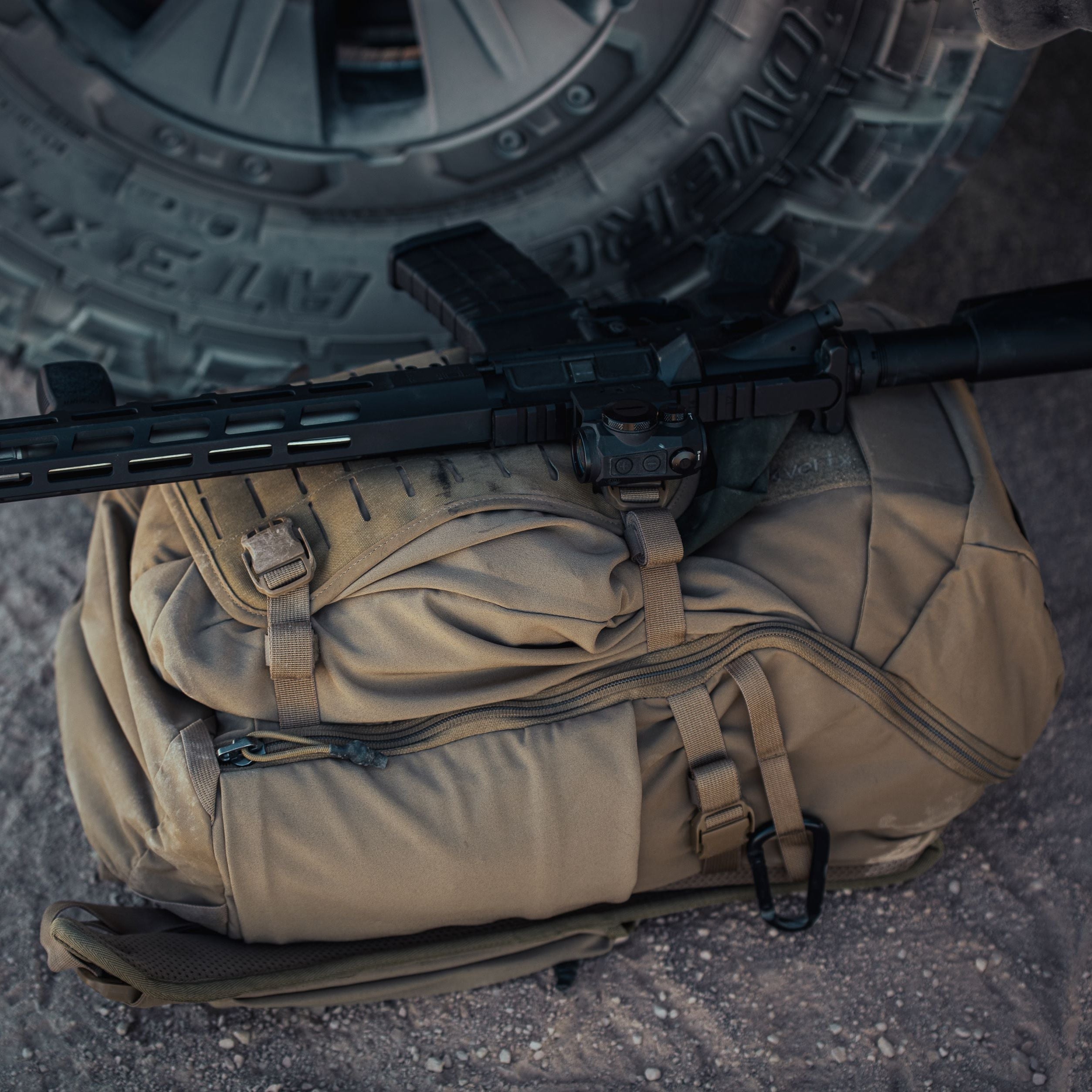 A black tactical rifle with a prominent red dot sight rests on a tan Vertx tactical backpack on dusty ground next to a BFGoodrich off-road vehicle tire