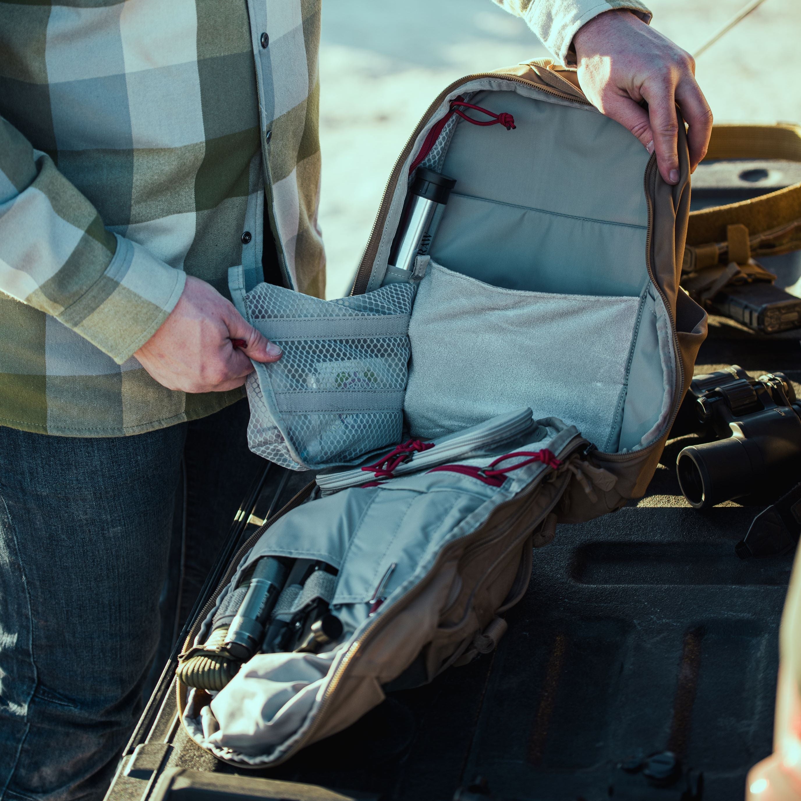 A person wearing a green and light blue plaid shirt organizes a khaki tactical backpack, revealing red cord loops, a silver item labeled RAW, and binoculars on a dark textured surface