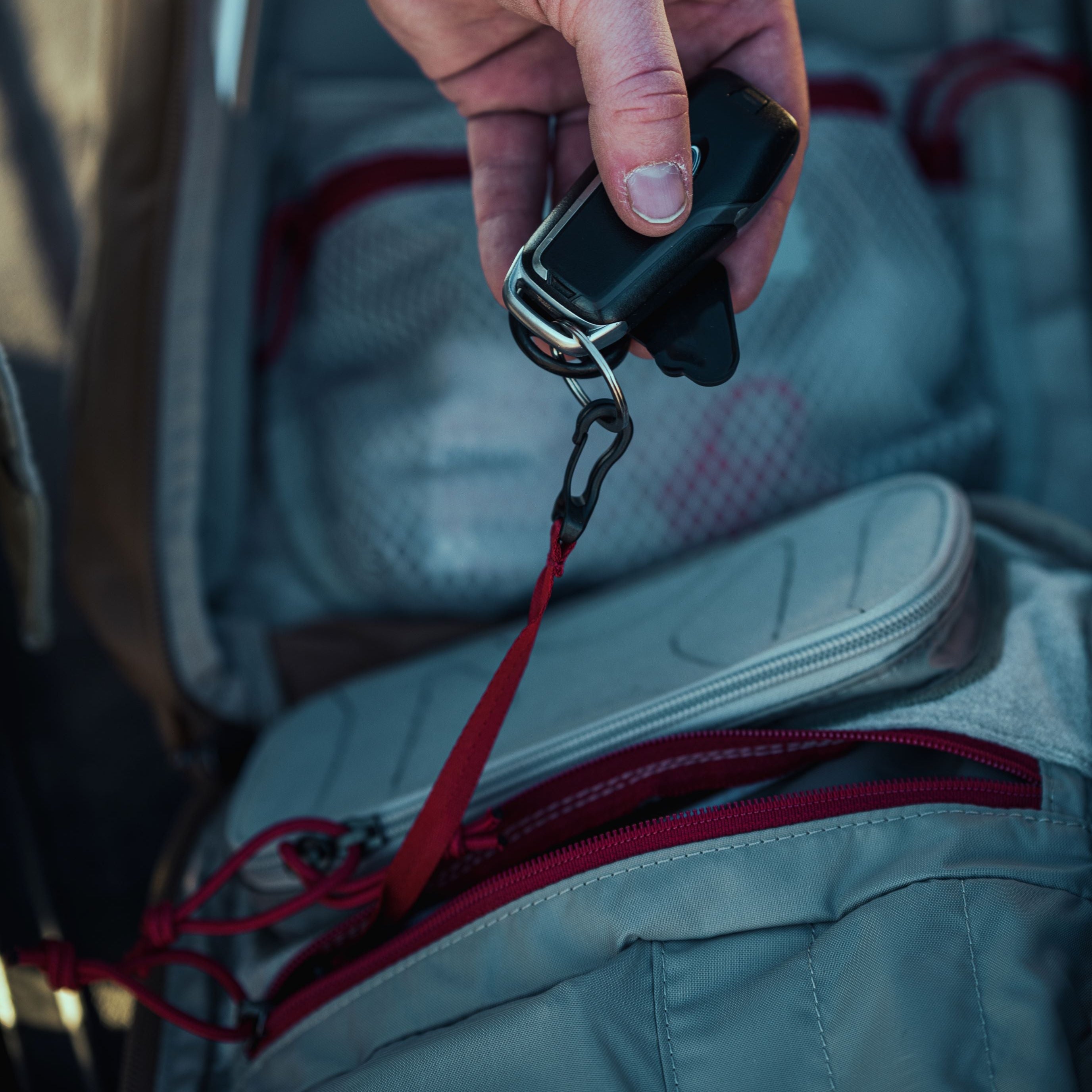 Close-up photograph of a hand holding a black car key fob with a red strap above an open grey backpack featuring bright red zippers and mesh pockets