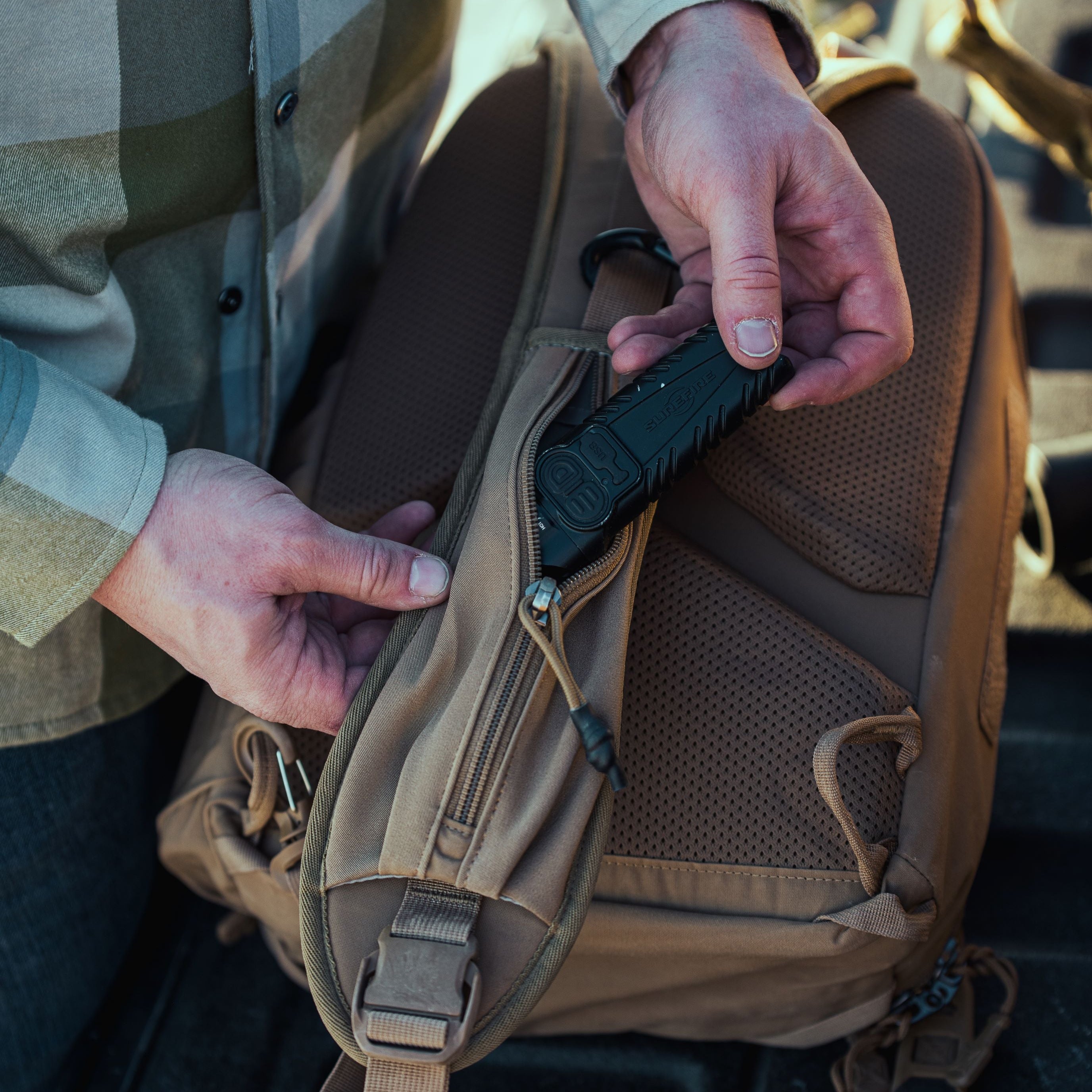 A person in a green plaid shirt places a black SureFire utility tool into the zippered side pocket of a coyote brown outdoor backpack with mesh padding