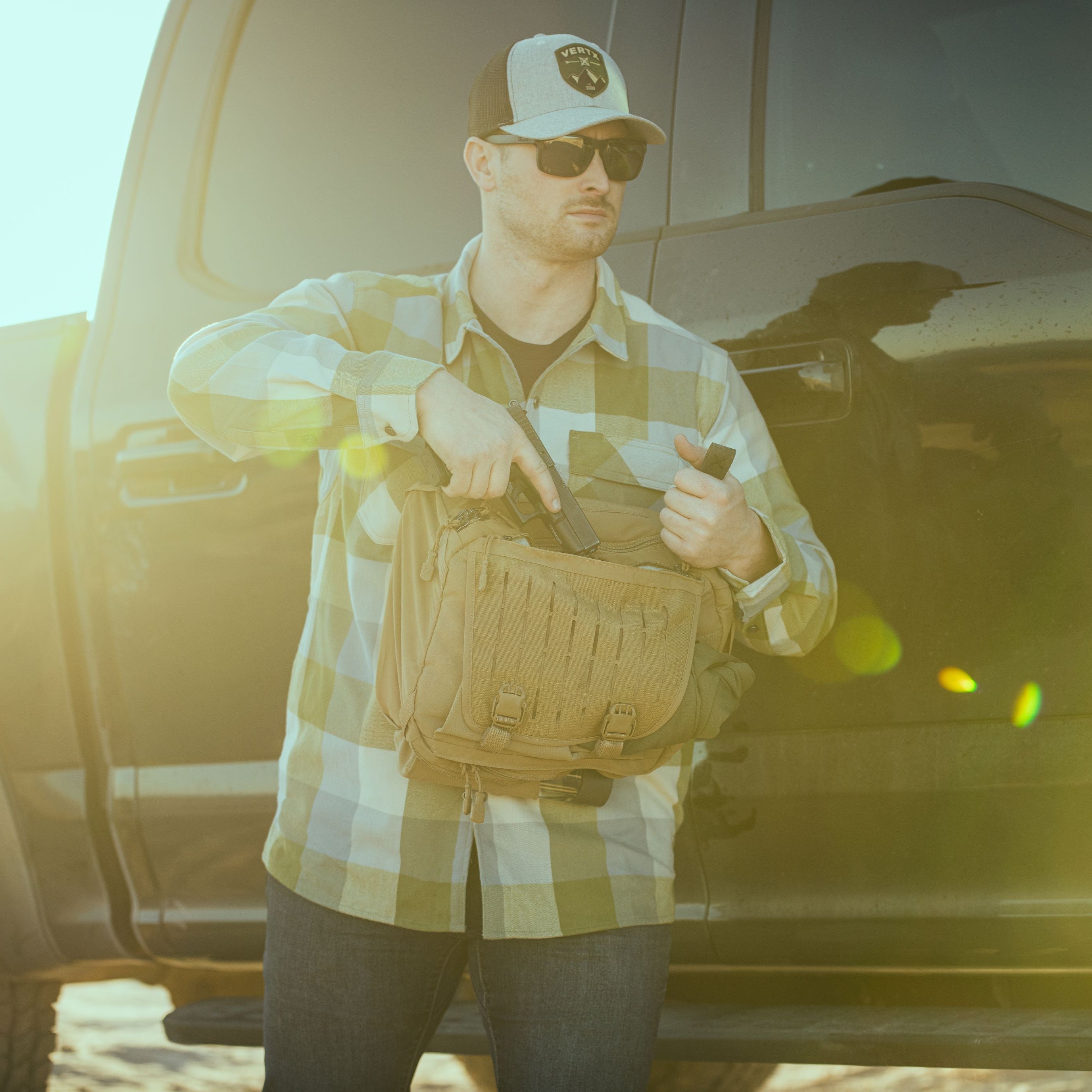 A man wearing a green and white plaid shirt, Vertx hat, and sunglasses inserts a black handgun into a tan tactical messenger bag while standing beside a dark pickup truck under a bright golden lens flare
