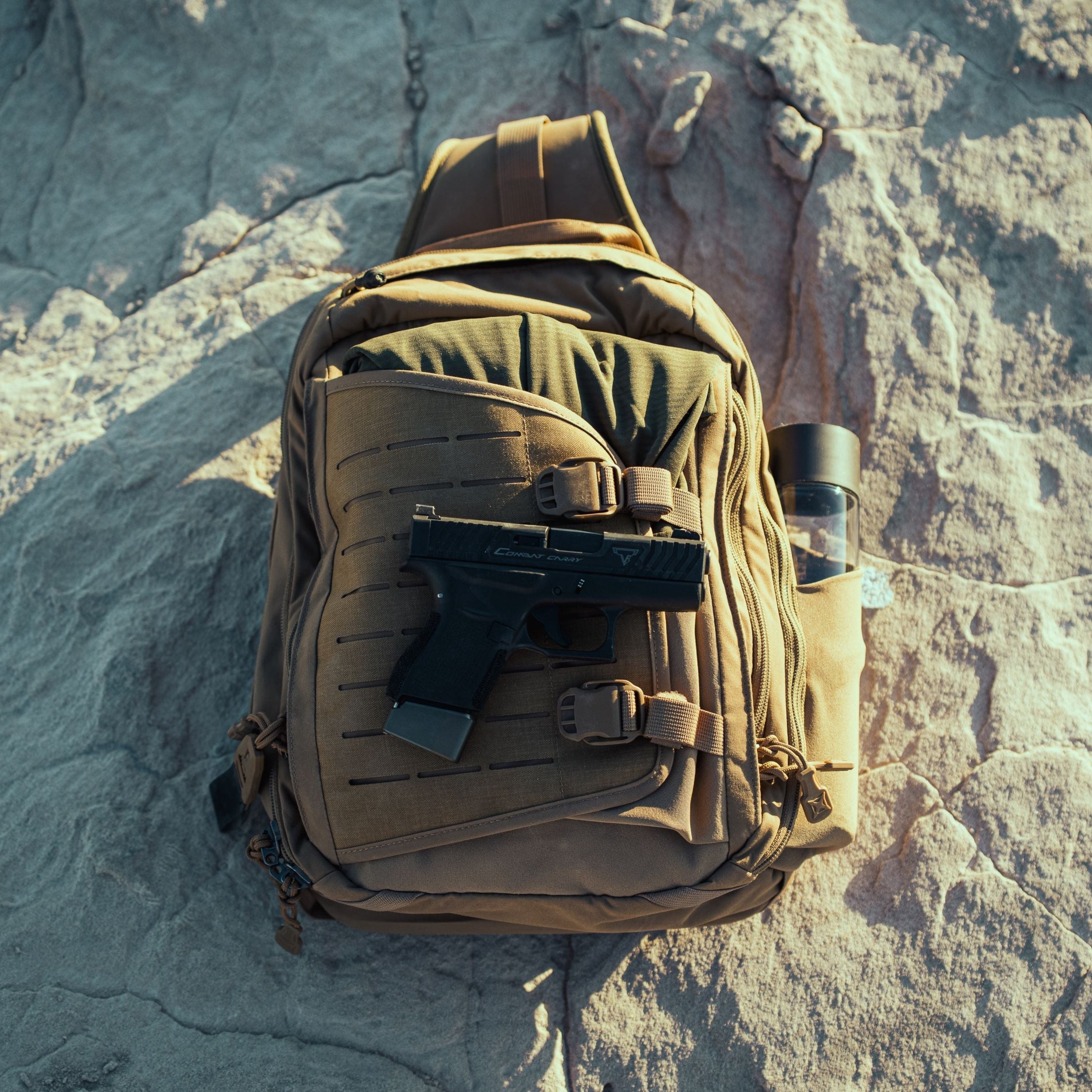 Photography of a tan tactical sling bag featuring MOLLE webbing, a black semi-automatic pistol with "Combat Carry" text and an extended magazine, and a clear water bottle, all resting on a rugged sunlit grey rock surface
