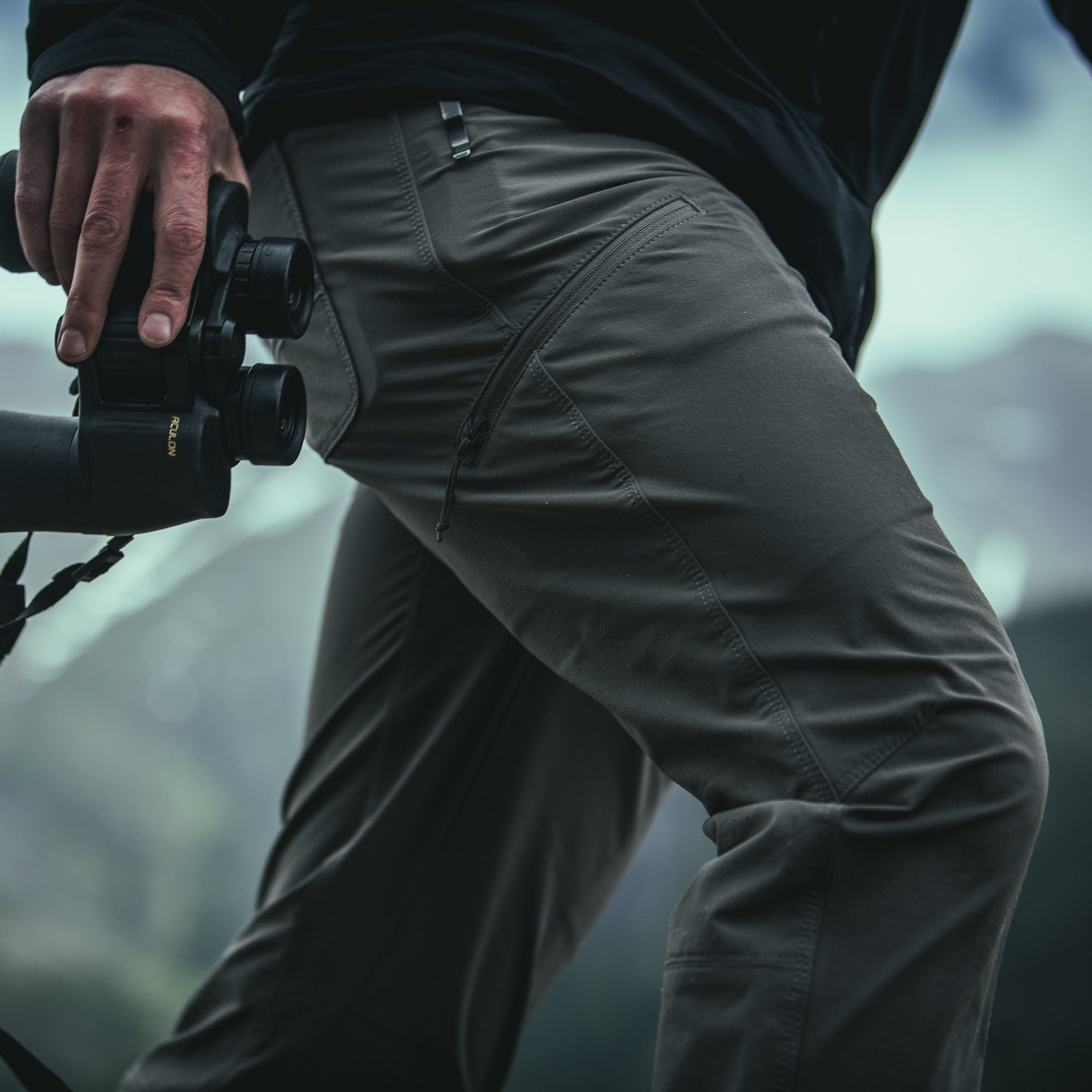 A photograph showing a person wearing dark gray hiking pants with zippered pockets holding binoculars against a backdrop of mountainous terrain featuring subtle blues and greens in the distance