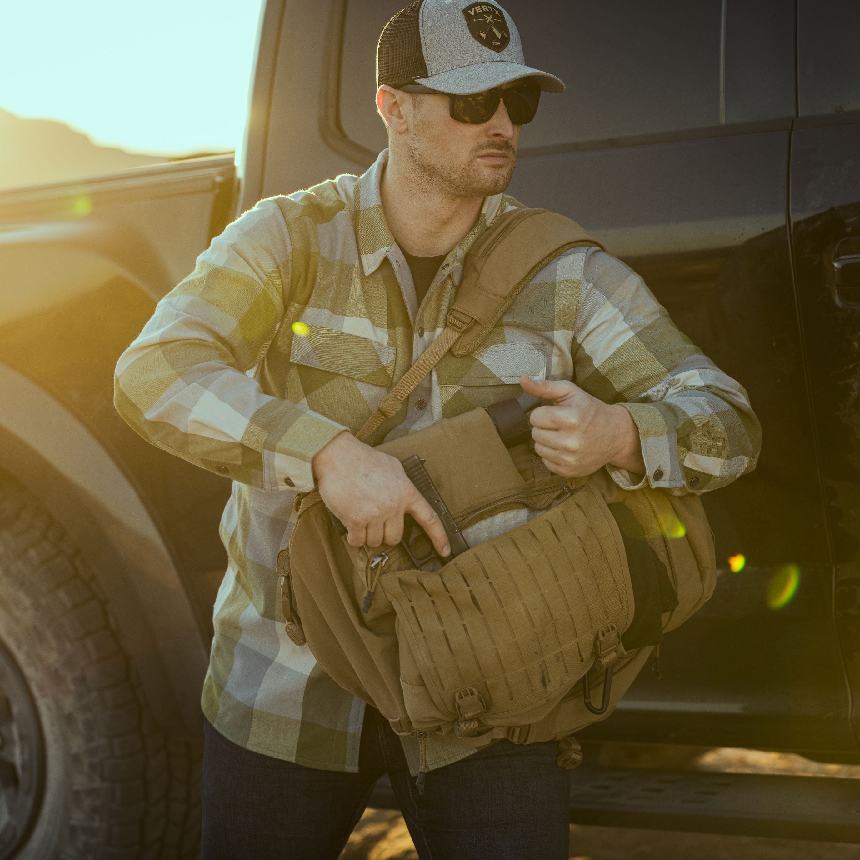 A man in a green and white plaid flannel shirt, sunglasses, and a grey Vertx cap drawing a black pistol from a tan tactical shoulder bag beside a dark truck in a sun-drenched desert at golden hour