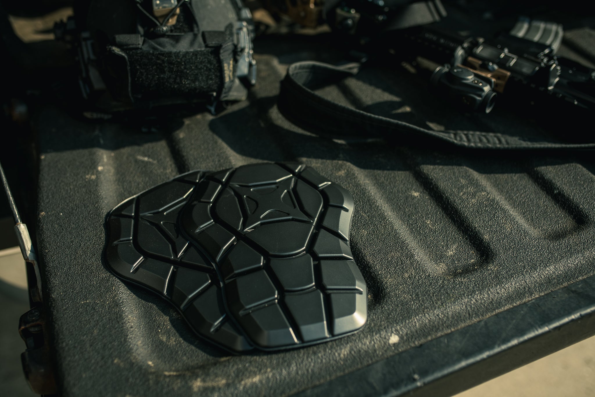 Photograph showing two black, textured, protective pads on a truck bed next to a rifle and tactical gear featuring a honeycomb pattern and glossy sheen