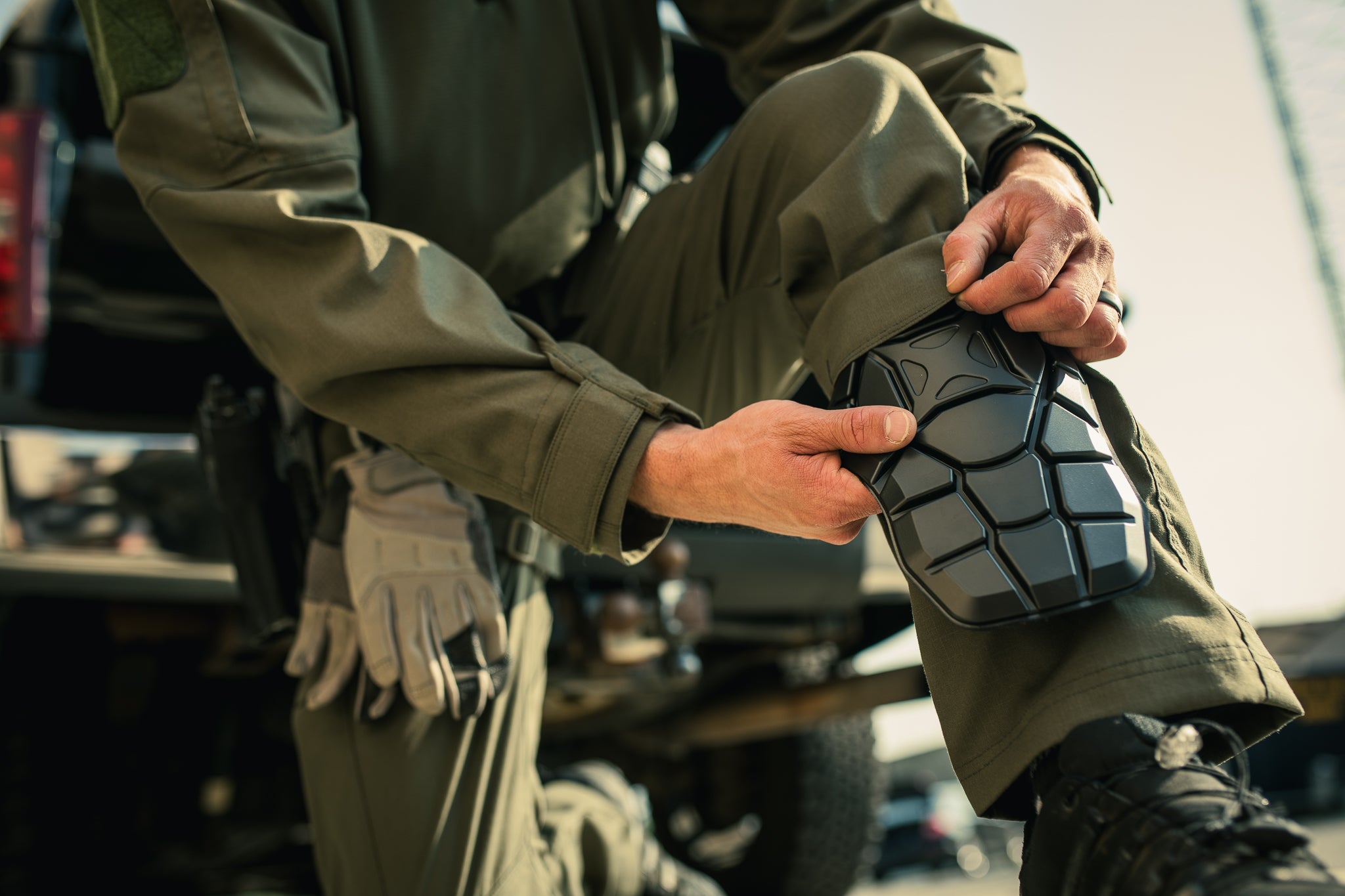 A photograph showing a person in olive green tactical pants and gloves attaching a black, geometrically patterned knee pad outdoors near a vehicle with a blurred background