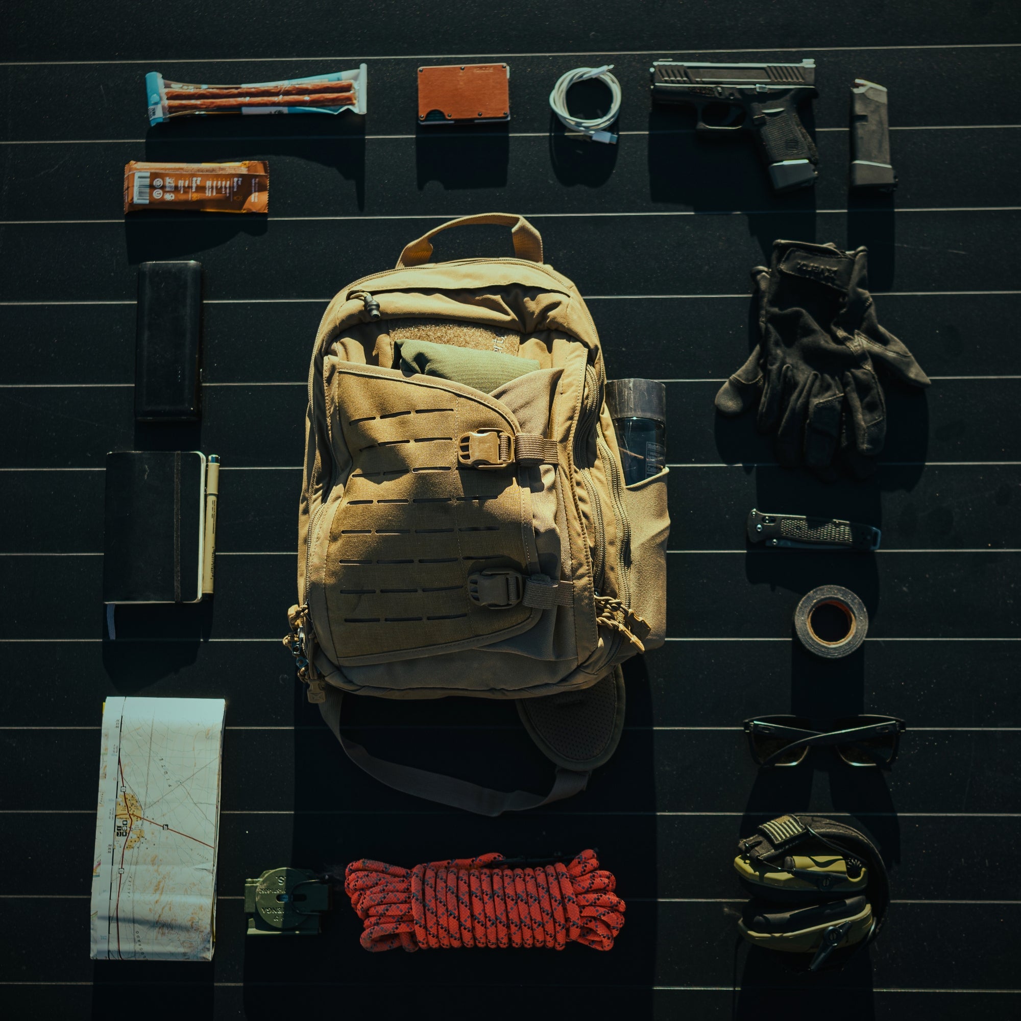 Photography uncredited a flat lay of survival and everyday carry gear arranged on a dark striped surface featuring a tan tactical backpack a black handgun with magazine and a bright orange climbing rope