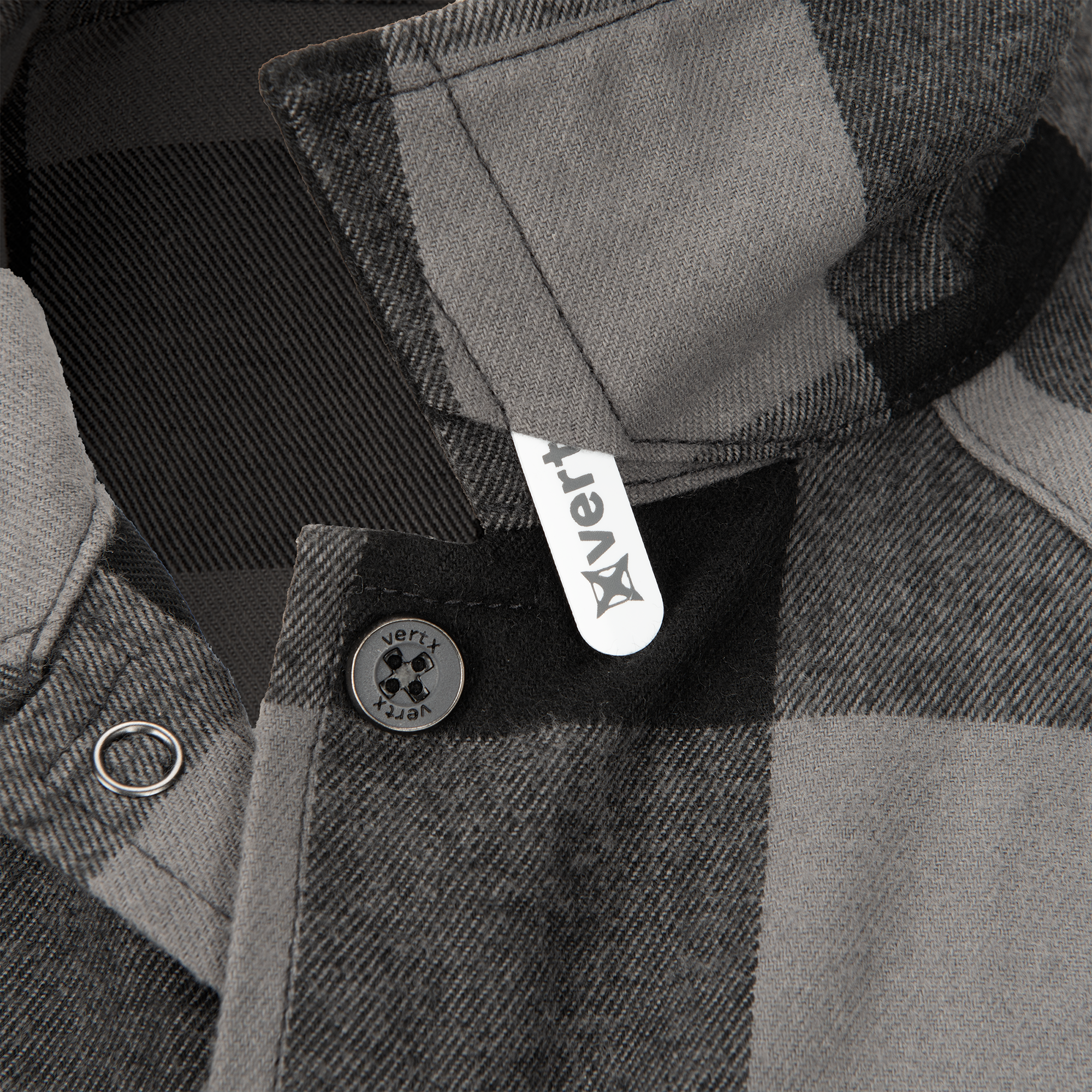 Product shot of a Vertx flannel shirt showcasing its gray and black plaid pattern, a metal button, and a white woven label with logo
