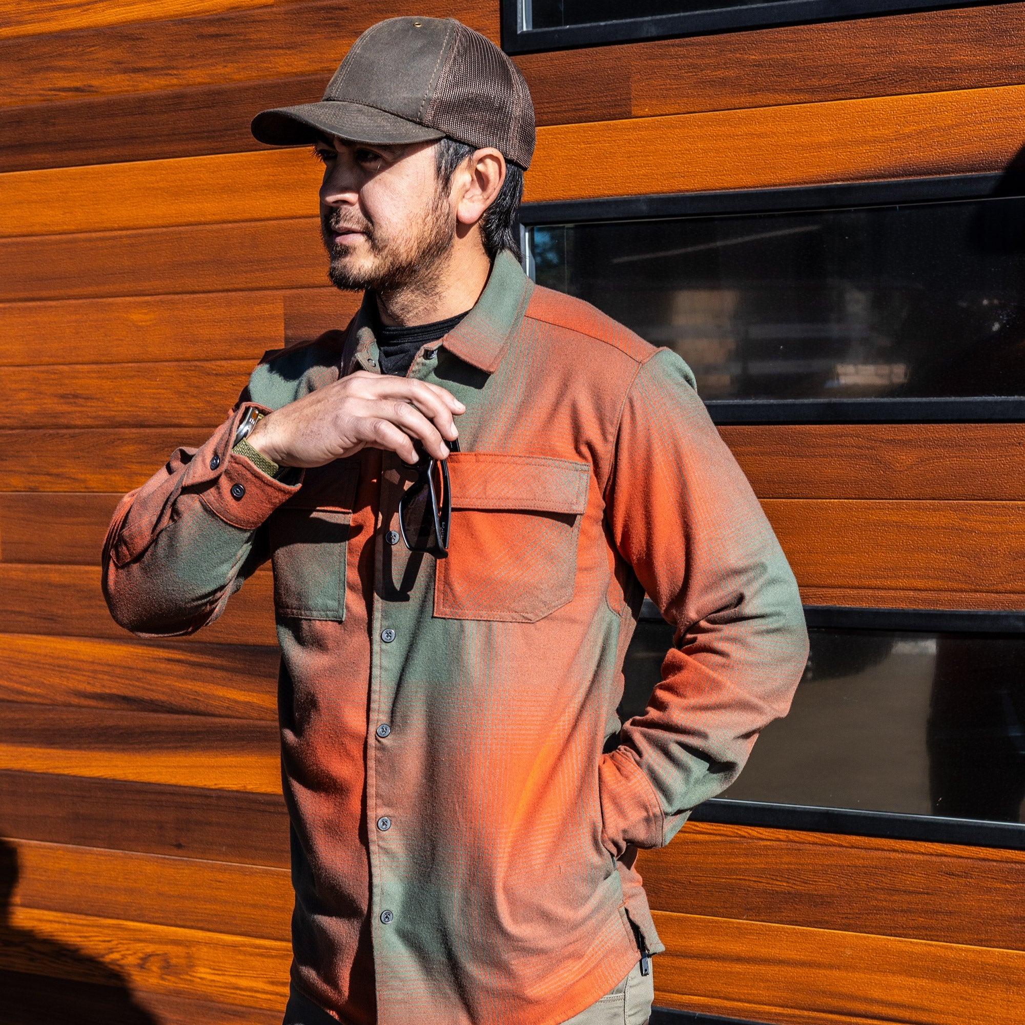 Photograph of a man wearing an orange and green plaid shirt near a rich-toned wood wall with black framed windows showcasing the shirt's textured fabric and the man's dark cap and sunglasses.