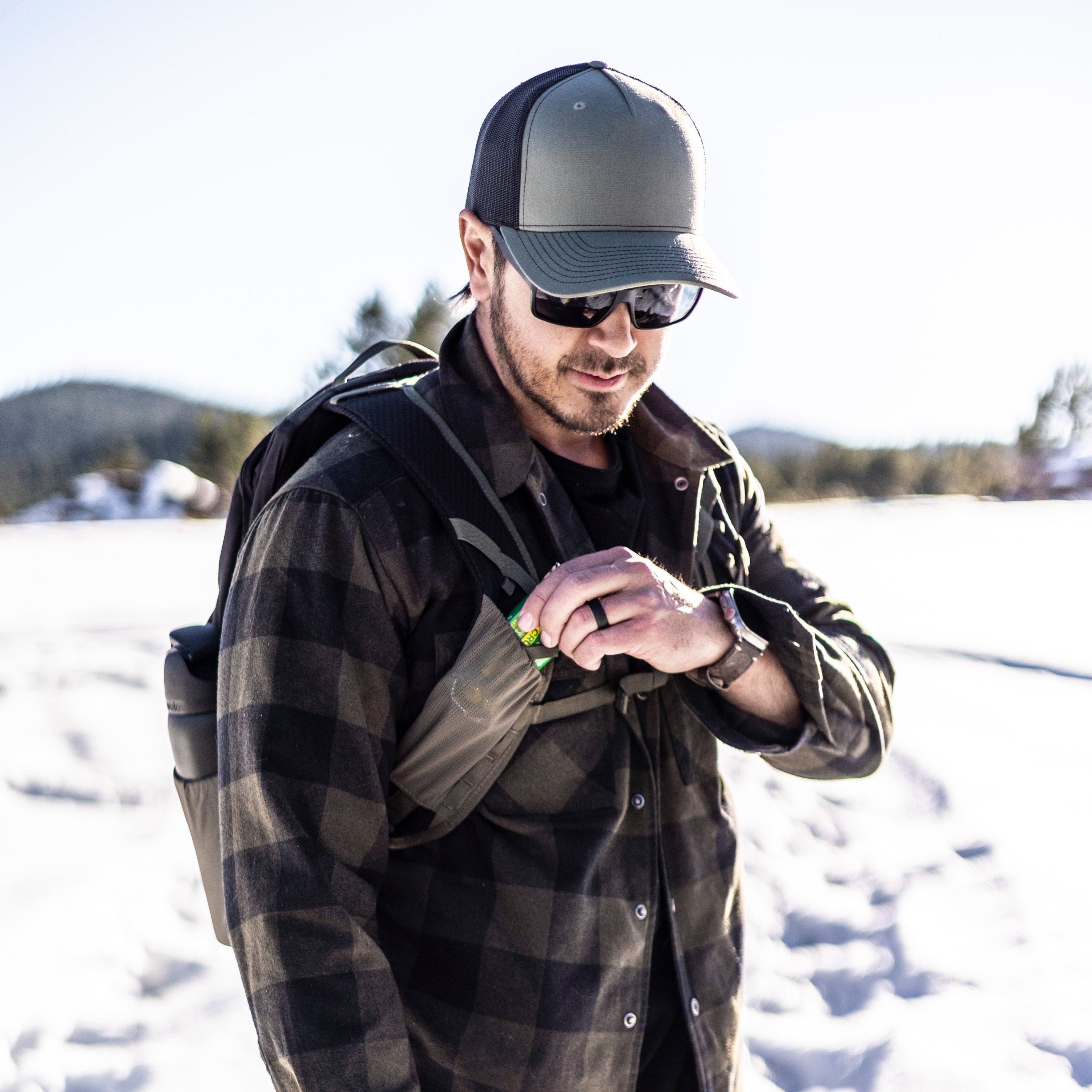 Photograph of a man in a grey and black flannel shirt, sunglasses, and a grey and black trucker hat accessing a green energy bar from his backpack in a snowy mountainous landscape with distant hills