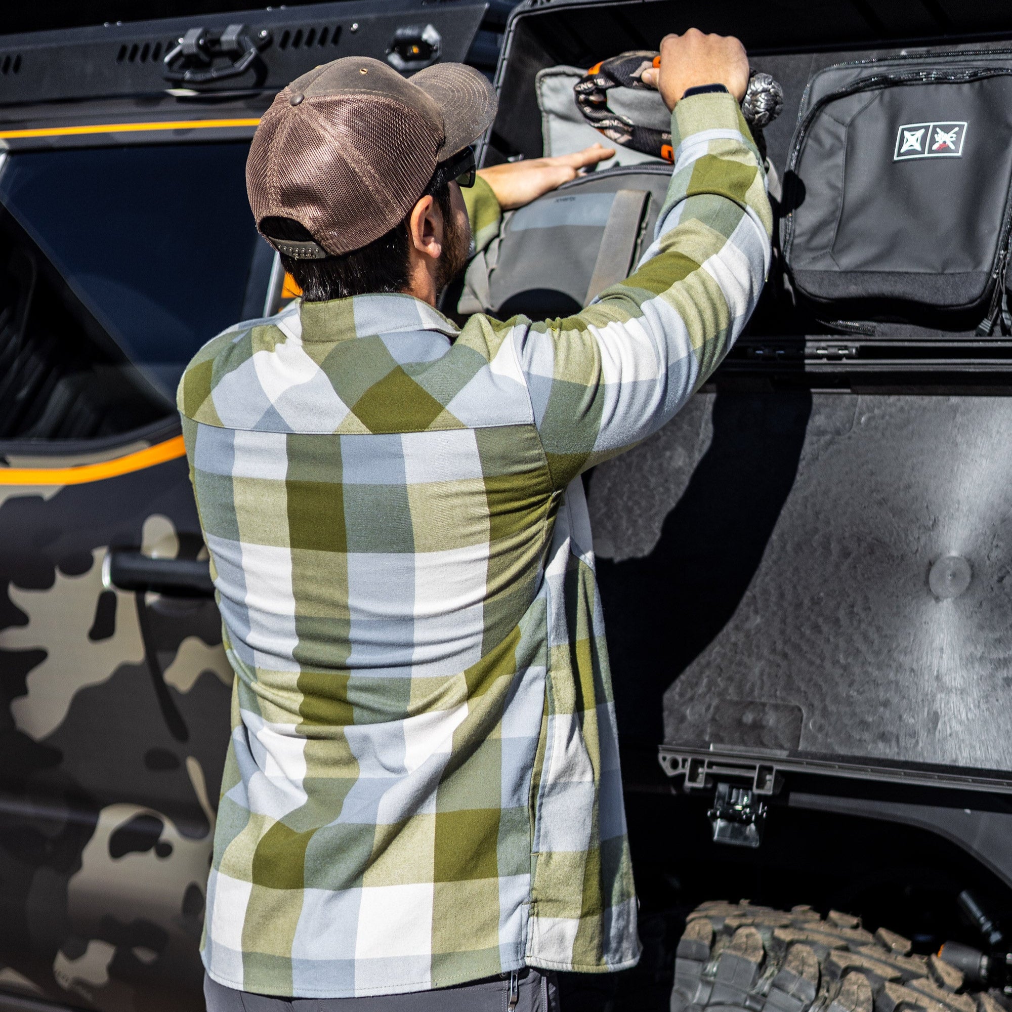 Photograph showing a person wearing a green and white plaid shirt packing grey and black bags into the back of a camouflage-painted vehicle with orange accents and large tires
