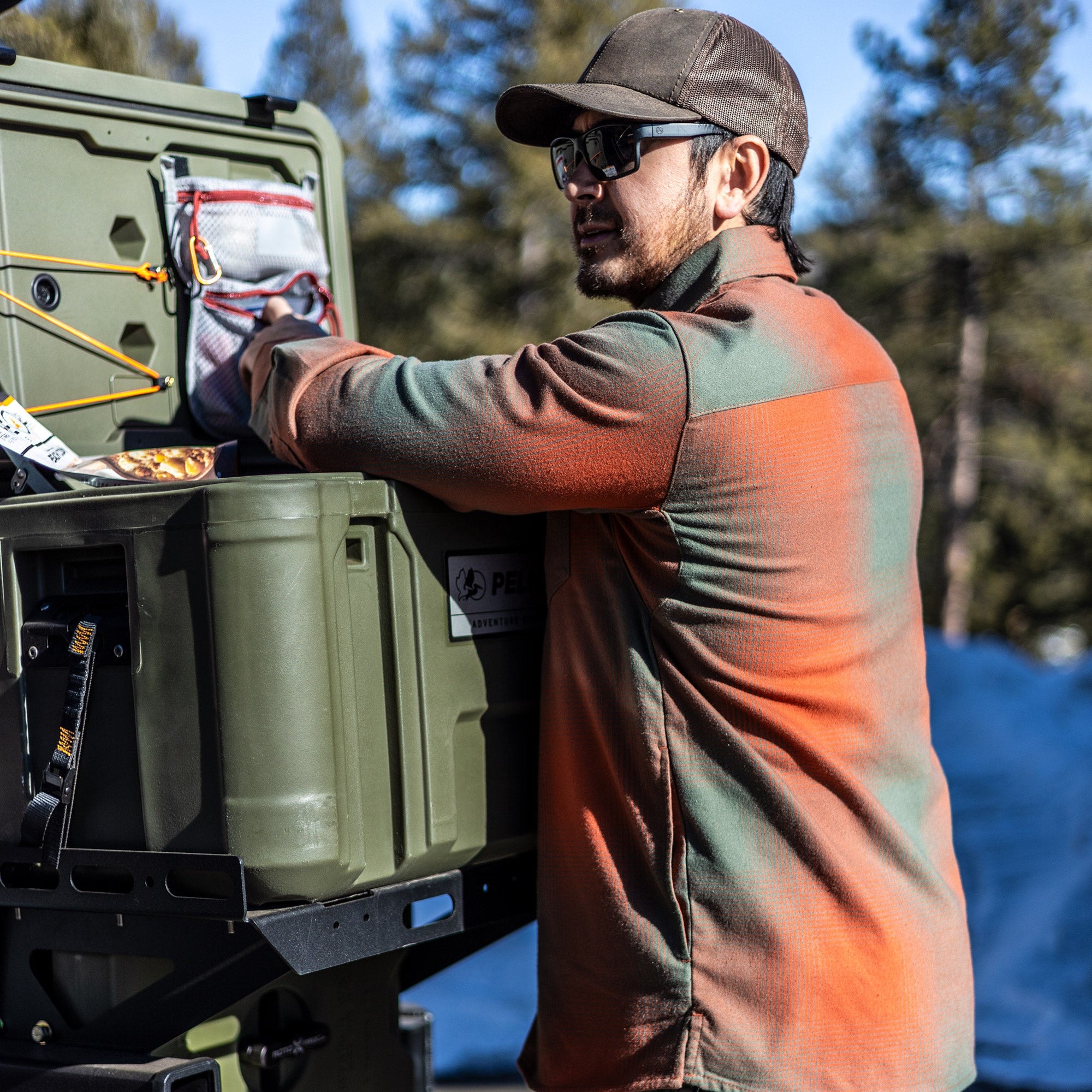 Photograph of a man in an orange and green plaid shirt packing a Pelican Adventure Gear cargo box outdoors near trees, featuring bright orange bungee cords, a gray mesh pouch, and a black metal rack.