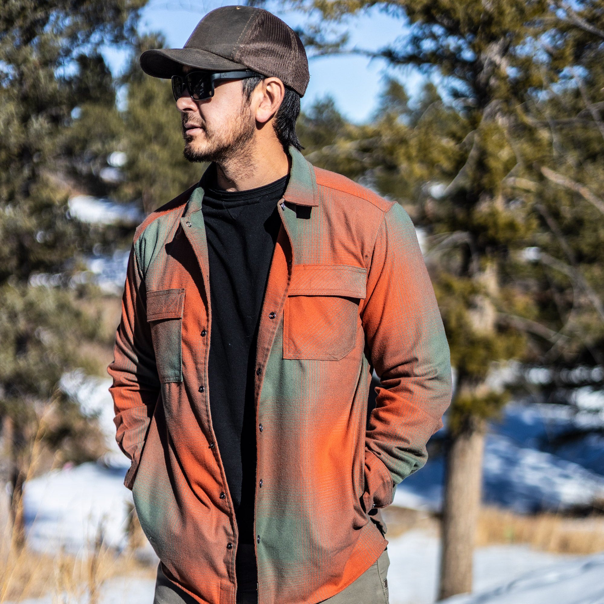 Photograph of a man wearing a burnt orange and sage green plaid shirt, sunglasses, and a brown cap standing in a snowy mountain landscape with tall evergreen trees