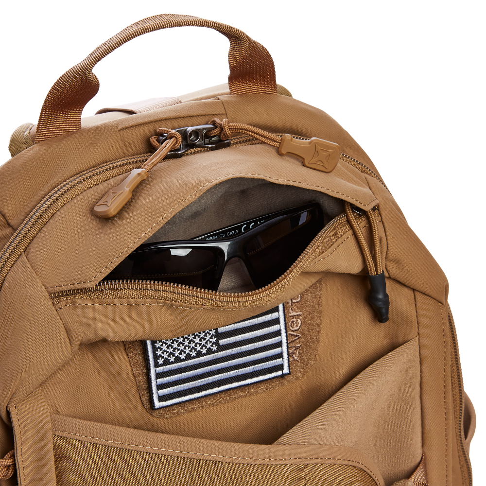 Close-up product photo of a tan tactical backpack with dark sunglasses in an open zippered pocket and a black and white American flag patch displaying Avert text