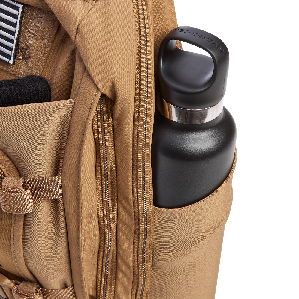 A tan tactical backpack with a black Hydro Flask water bottle in its side pocket, displaying robust zippers and a black and white American flag patch
