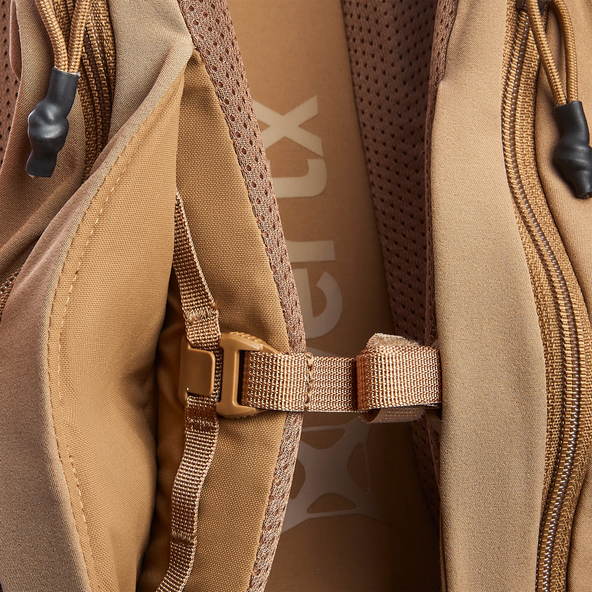 Arc'teryx product photography showing a close-up of a tan backpack with a visible brand logo, rugged fabric texture, and a tan plastic buckle