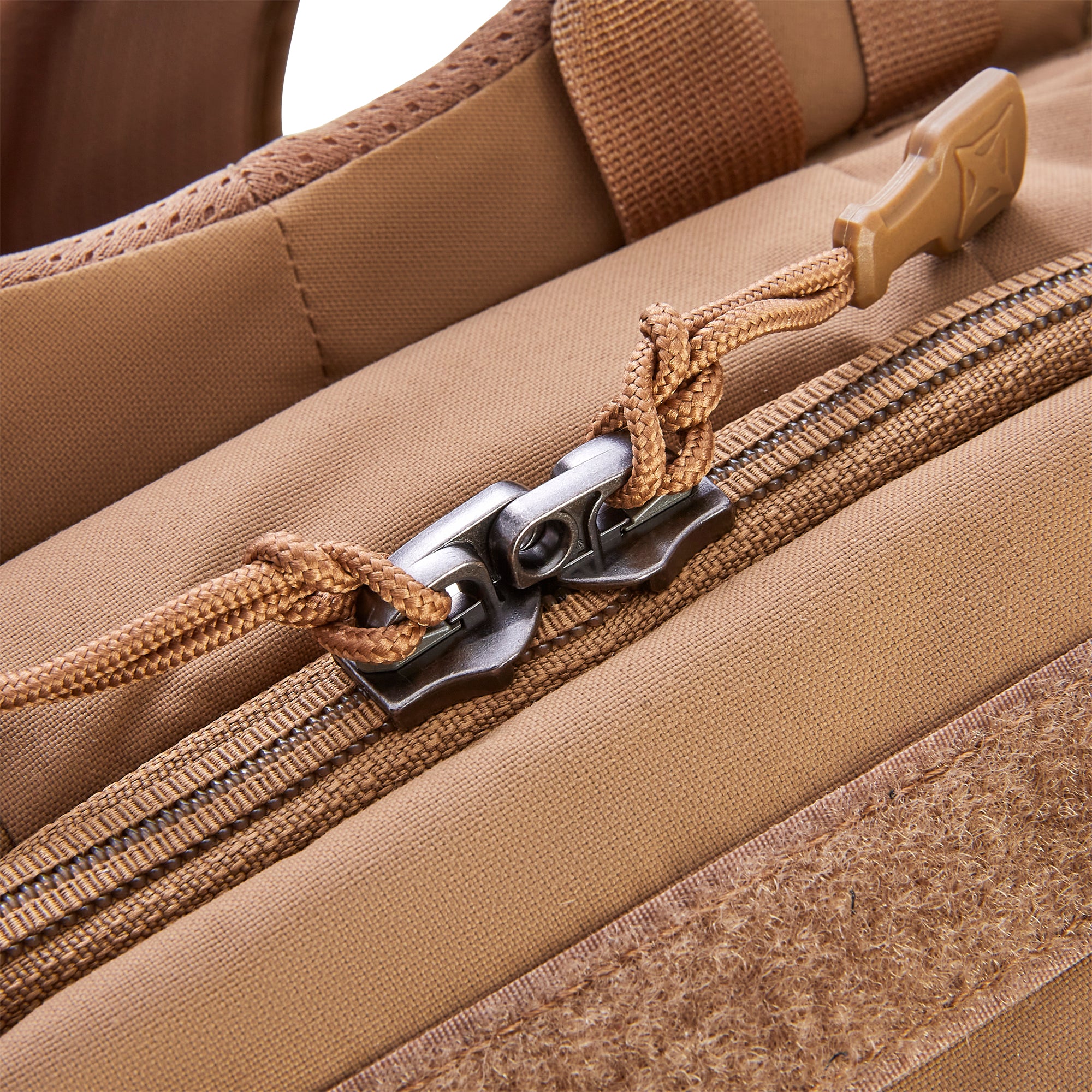 Close-up of a tan tactical bag showing dual dark metal zipper sliders with braided tan paracord pull tabs, a distinctive tan plastic pull tab with an embossed logo, and a brown hook-and-loop Velcro patch on durable canvas-like fabric