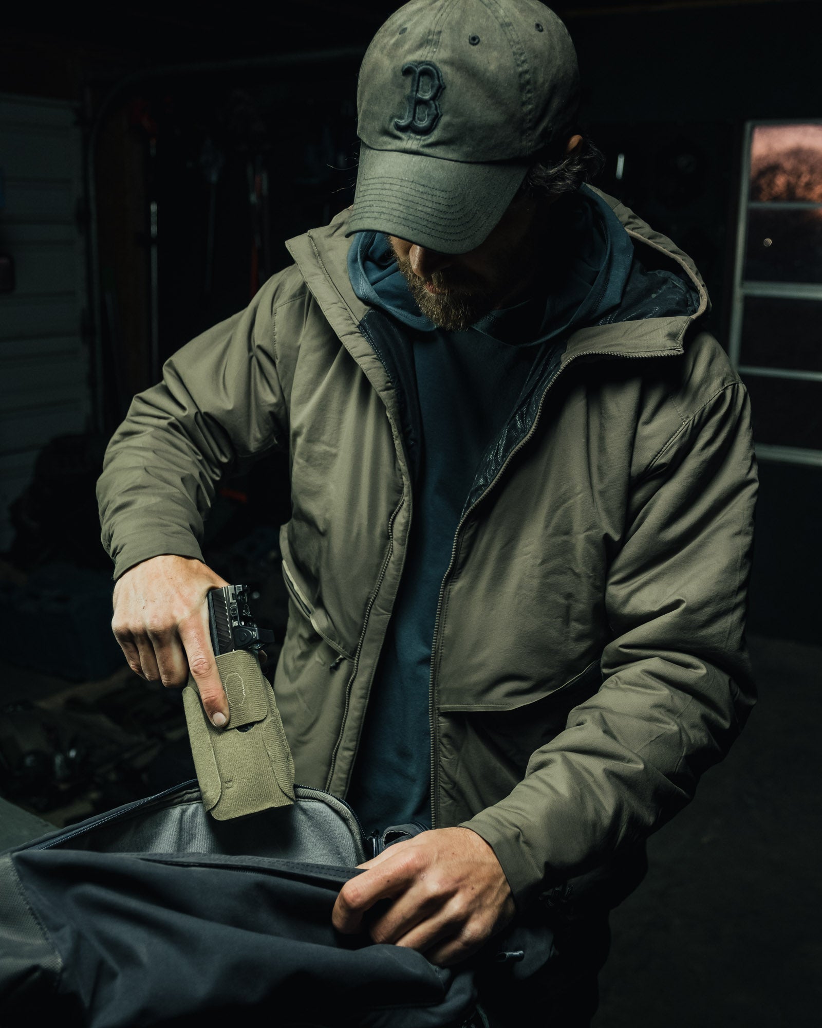Photography, unknown artist: A man in a dark olive green jacket and Boston Red Sox cap packs a dark handgun into a muted green case inside a black backpack in a dimly lit garage with visible tools and a window showing a twilight sky