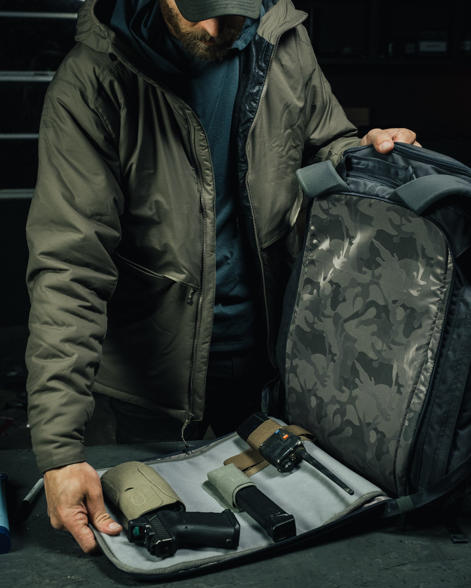 Photography a man in a olive green jacket unpacks a tactical backpack revealing a handgun, magazine, and radio with tan and grey accents in a dark setting