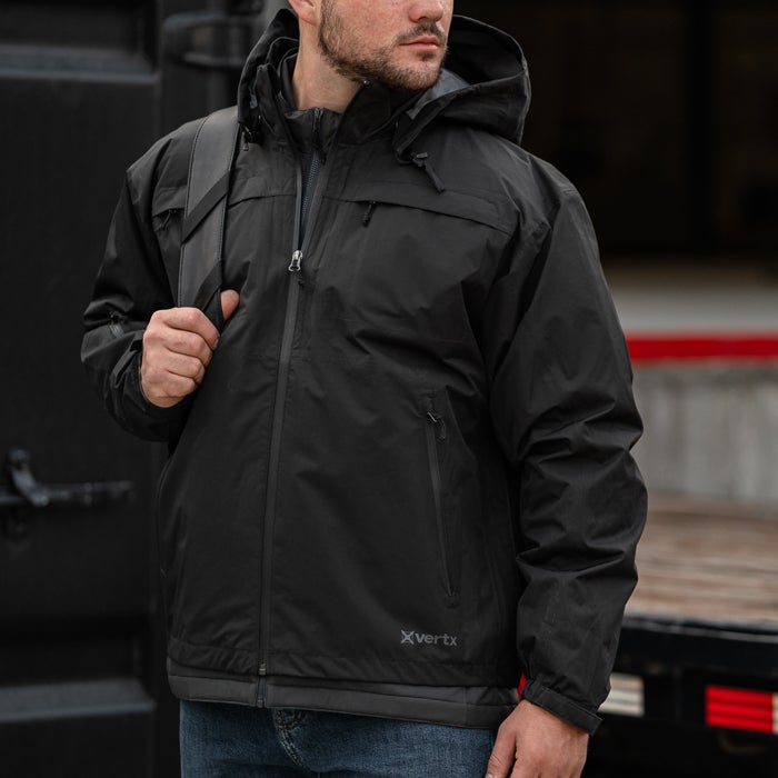Product photography showing a man wearing a black Vertex brand jacket with zippered pockets and a gray inner lining outdoors near a dark truck and red accents