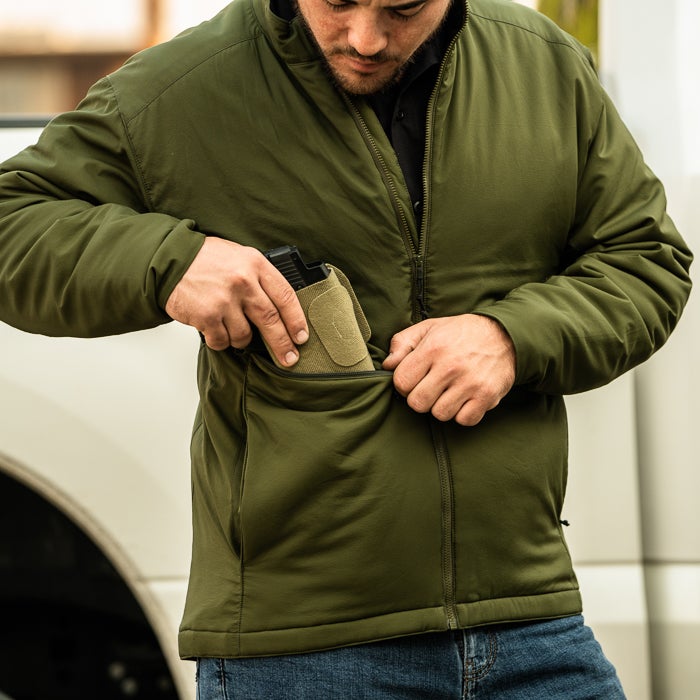 Photograph of a man in an olive green jacket concealing a handgun in a tan pocket liner against a blurry urban backdrop