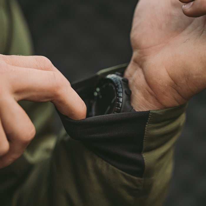 Close-up photograph showing a person's hand adjusting the cuff of an olive green and black long-sleeved shirt with a dark-colored watch visible on their wrist featuring ribbed detailing and a contrasting fabric panel