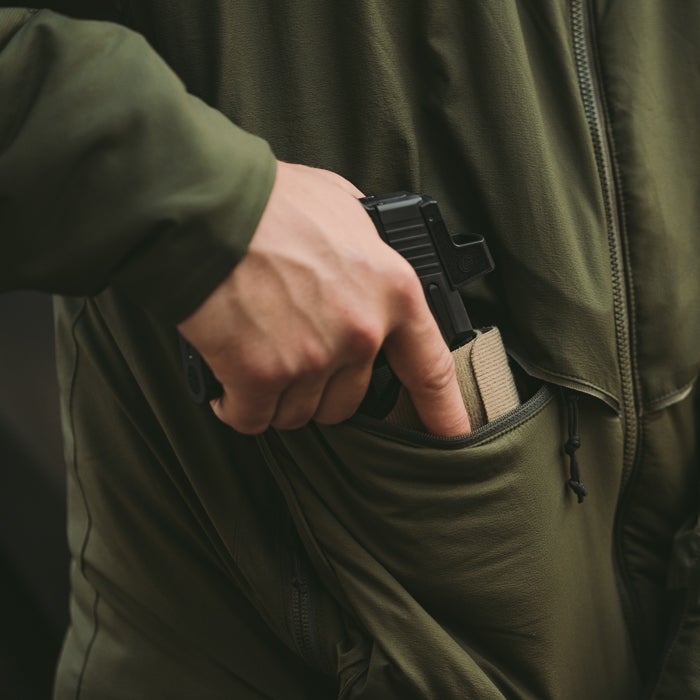Photograph showing a hand inserting a black handgun into a tan holster in an olive-green jacket pocket, the fabric texture is visible, the zipper is partially visible, and the lighting is muted.