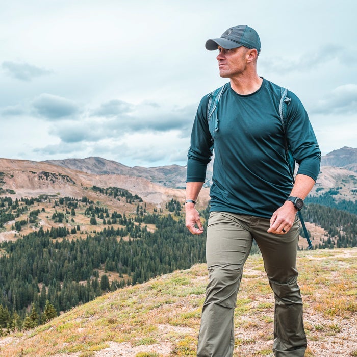 Photograph of a man hiking in the Colorado mountains showcasing a dark teal shirt olive pants and a smartwatch against a backdrop of rolling hills and evergreen trees