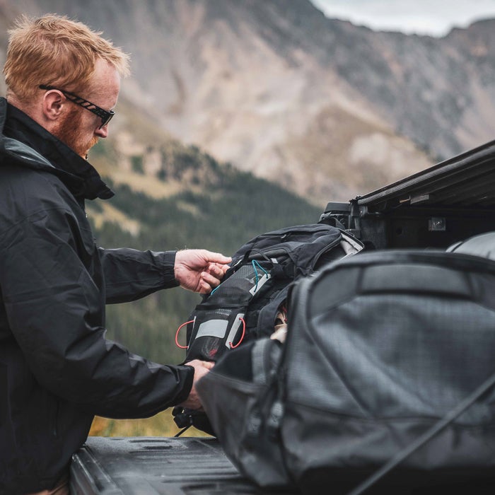 Photograph of a man packing black backpacks with red and gray accents into a truck bed against a mountainous backdrop featuring textured rock formations and lush greenery
