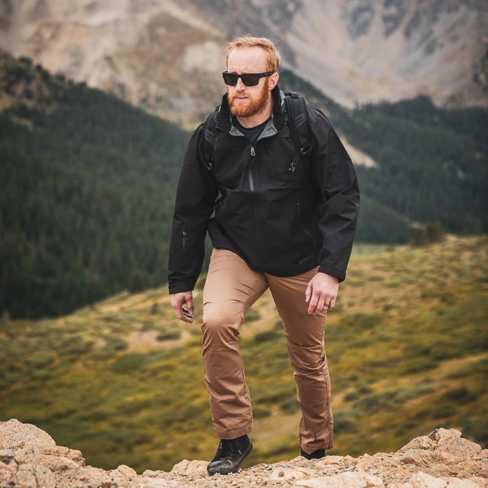 Photography of a man in a black jacket and khaki pants hiking a mountain range with dark green pine trees and rocky outcrops in the background showcasing his stylish sunglasses and beard
