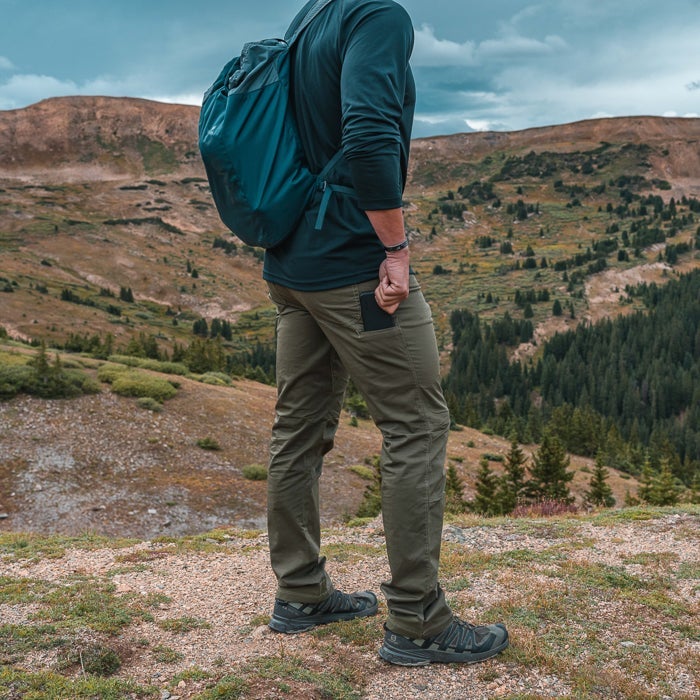 Photography A hiker with a teal backpack stands on a Colorado mountain overlooking a valley of evergreen trees and ochre hills a dark gray phone in his pocket and olive green hiking pants