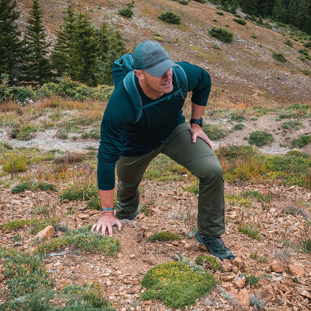 Photograph of a man hiking a rocky mountain trail wearing a dark green shirt, olive pants, and a gray American flag hat amidst low-lying shrubs and sparse trees