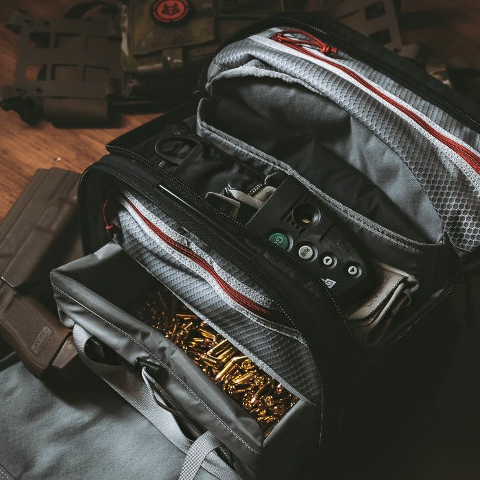 Photograph of a black and gray tactical range bag containing brass ammunition, a pistol, and other accessories with red zippers and gray mesh pockets