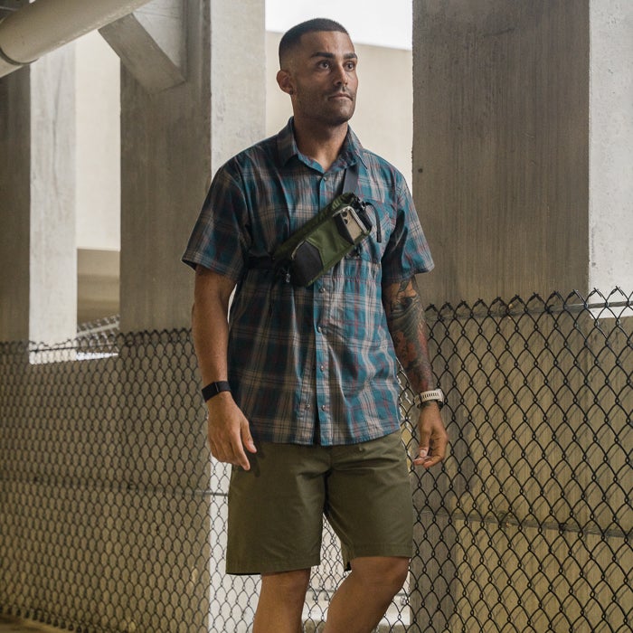 Photograph a man wearing a teal plaid shirt olive shorts and a green fanny pack standing in a parking garage with chain link fence and concrete pillars