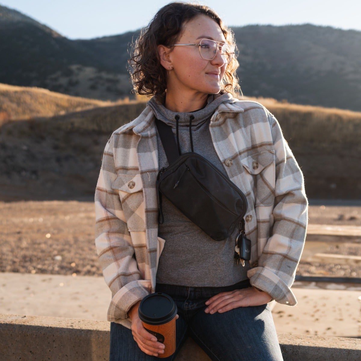 Photography Unknown Artist Woman in cream plaid jacket and glasses sits outdoors near mountains holding coffee a black fanny pack with keys attached a warm sunny day