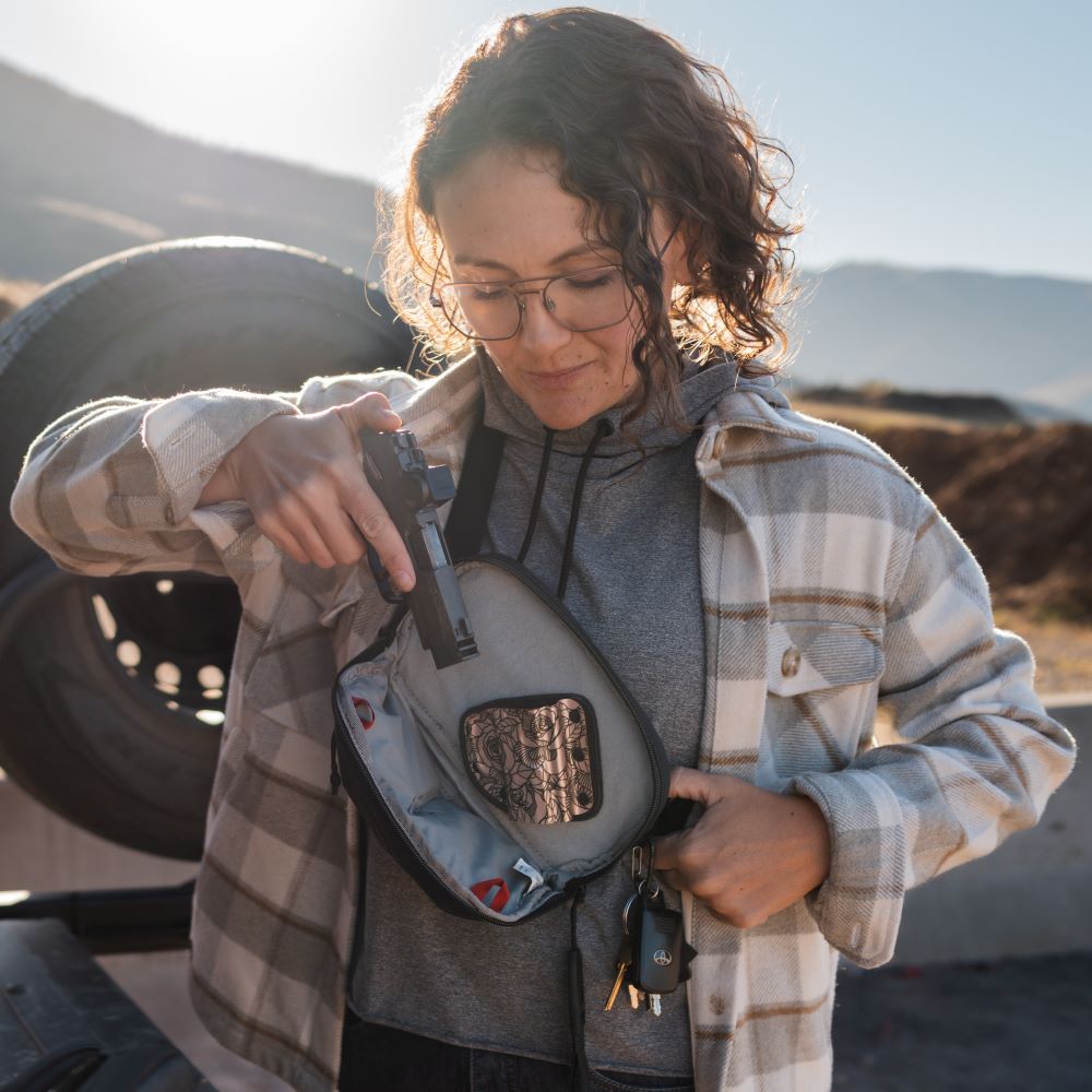 Photograph woman placing handgun into concealed carry bag outdoors near mountains and a tire with rose-patterned interior and keys visible.