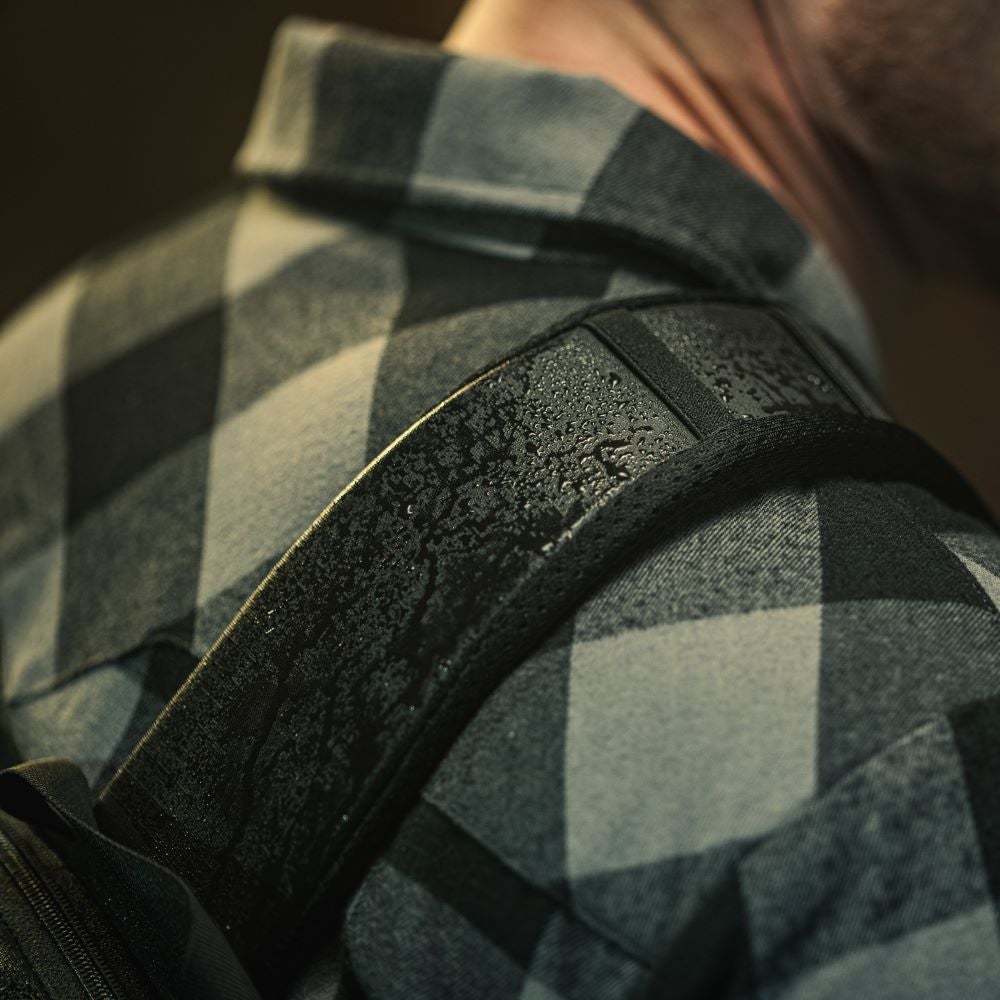 Close-up photograph showing a water-resistant backpack strap resting on a gray and black plaid shirt the strap displays numerous water droplets and a textured surface