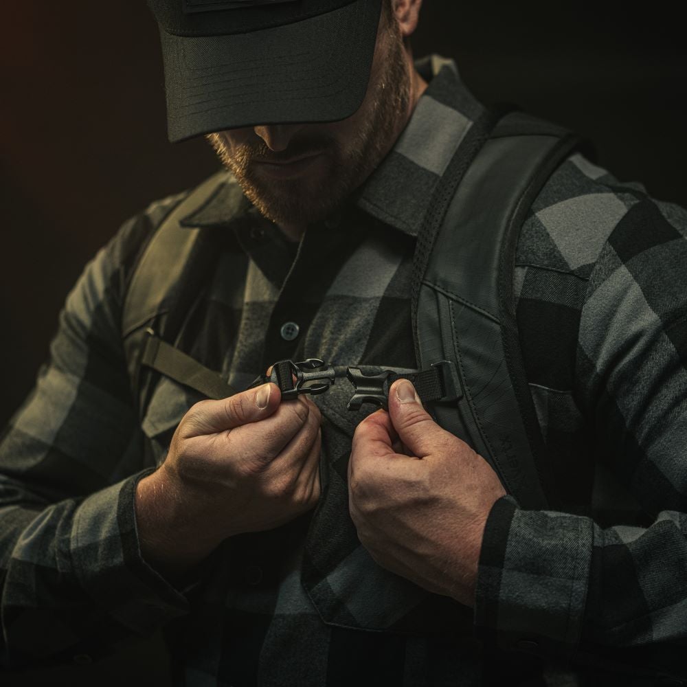 A photograph of a man adjusting a black backpack strap with a dark gray and black plaid shirt, strong hands and a black baseball cap.