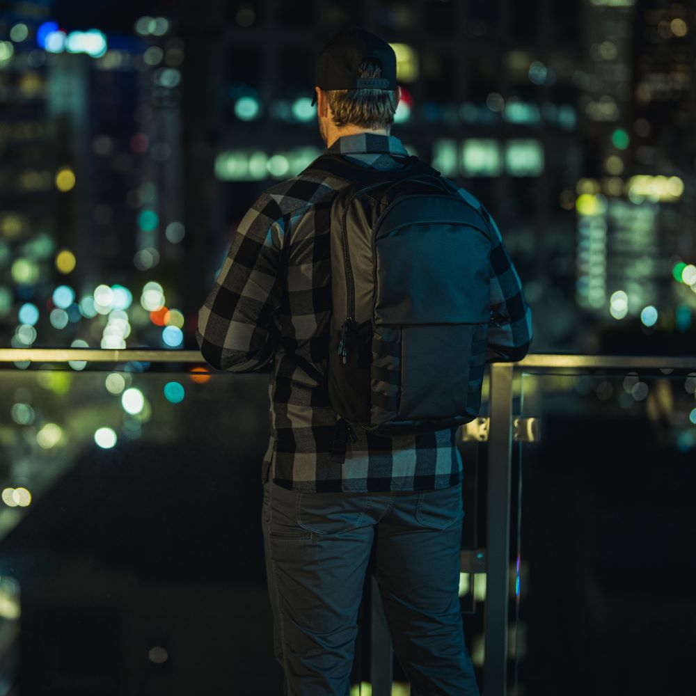 Night photography of a man with a black backpack on a city balcony overlooking blurred city lights and skyscrapers wearing a gray and black plaid shirt and gray pants