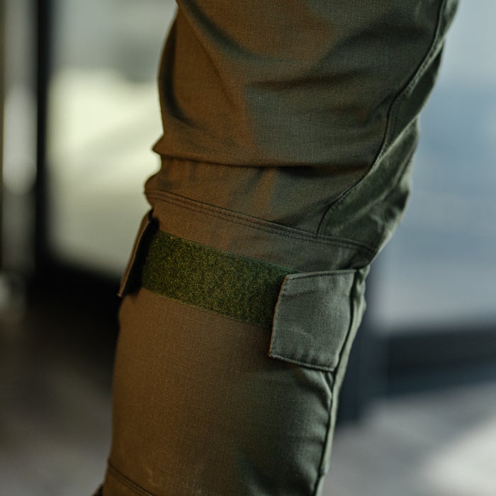 Close-up product shot of olive green tactical pants featuring a dark green Velcro strap knee pad and subtle stitching details showing the texture of the fabric