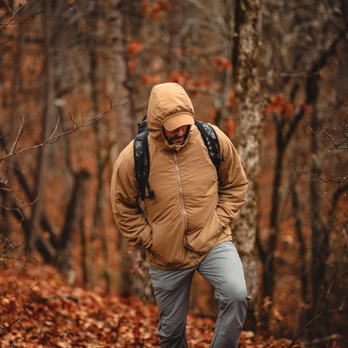 Photograph of a man in a tan jacket and gray pants hiking through an autumnal forest with bare trees and a carpet of reddish-brown leaves featuring a camouflage backpack and a baseball cap under the hood