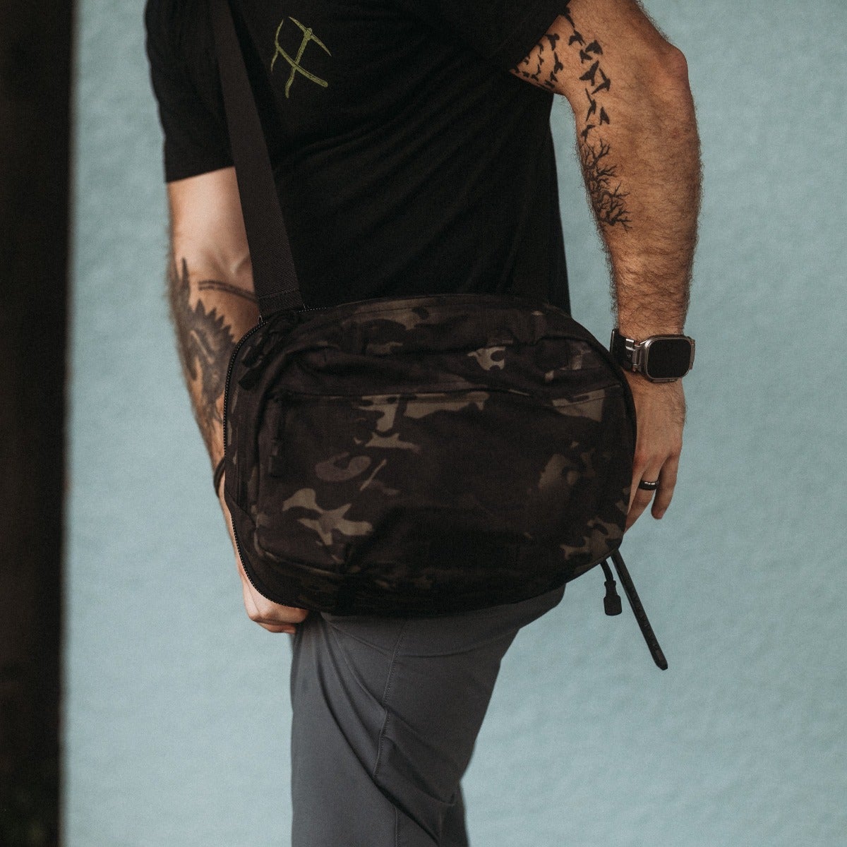 Photography Unknown Artist A man models a black camo crossbody bag against a pale blue background showcasing a watch a ring and a unique tattoo