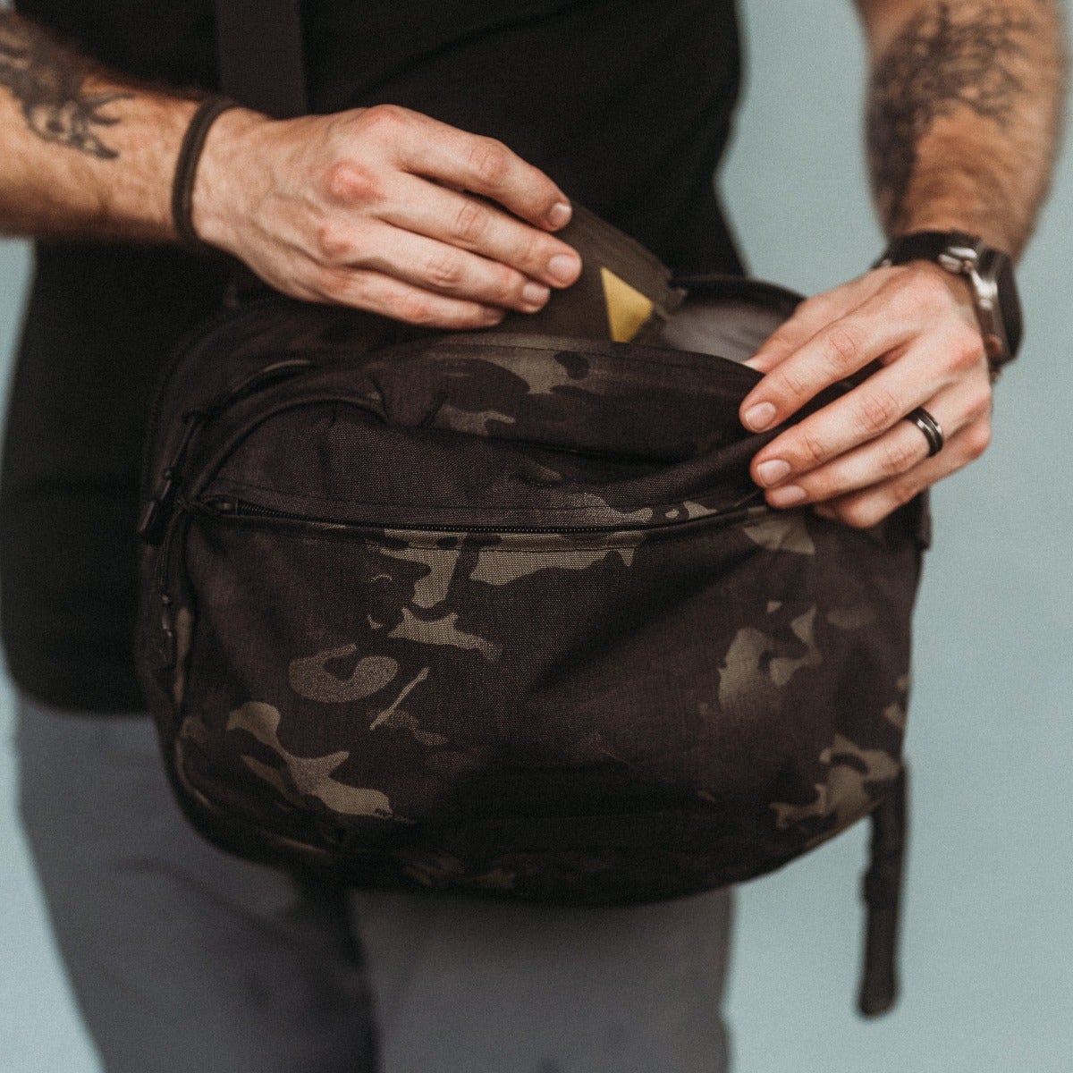 Photograph of a person's hands accessing a dark camouflage sling bag with a black zipper and olive-green accents showing a partially visible yellow item inside.